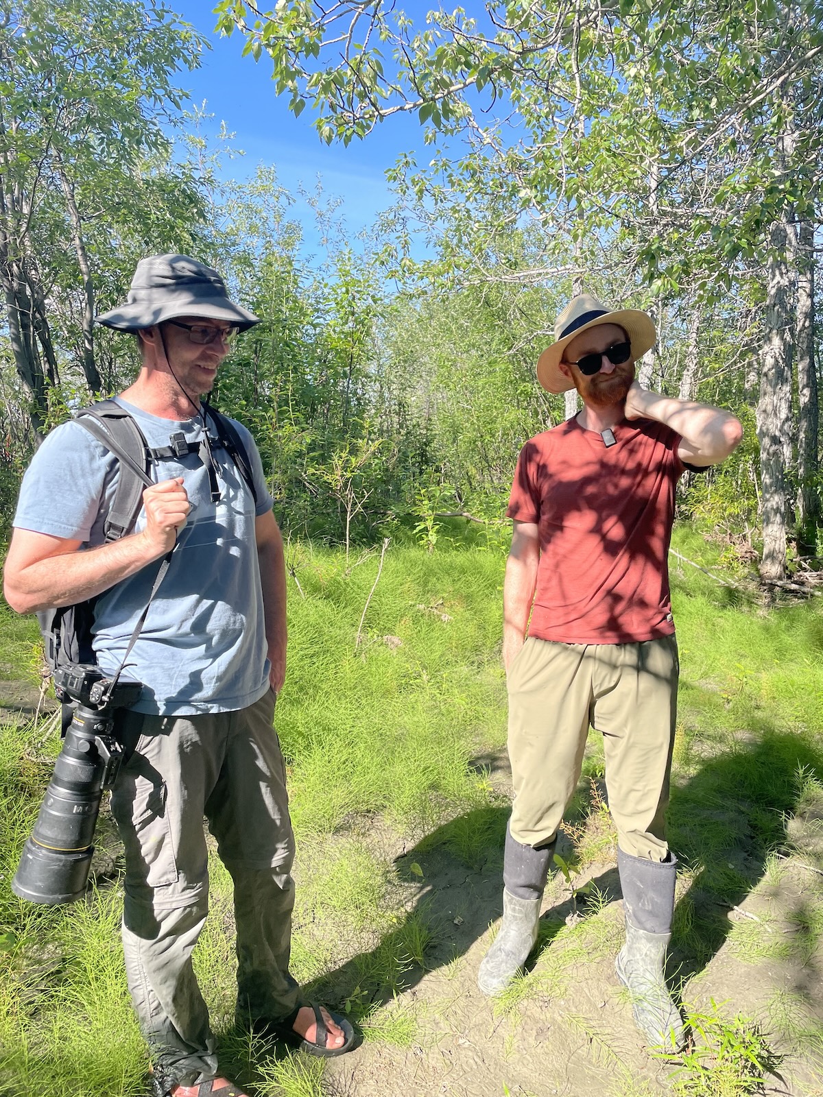 Two men wearing hats stand in dappled sunlight on a grassy area within a grove of deciduous trees. The man on the left carries a camera with a large telephoto lens.