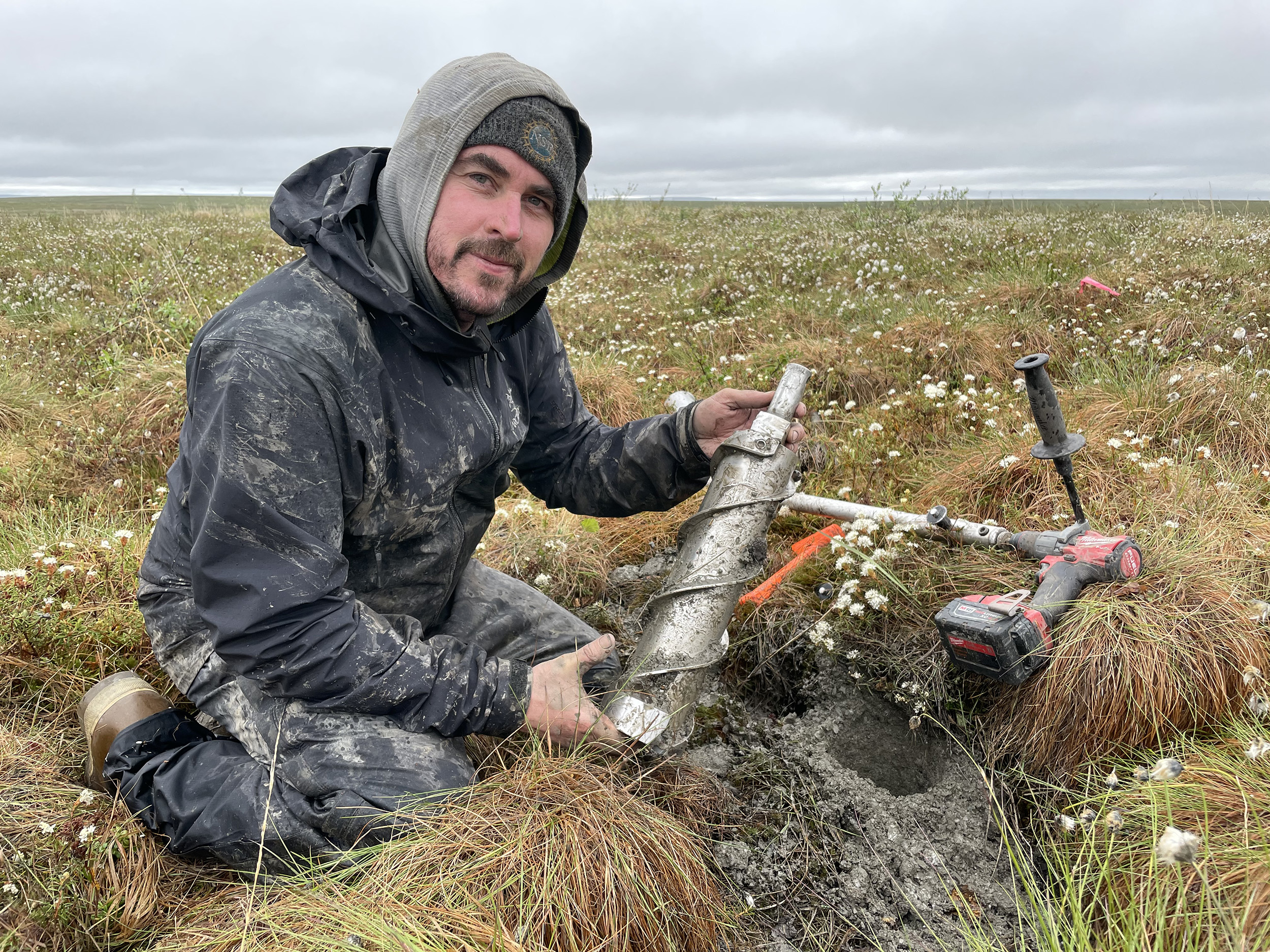 A man holds a piece of coring equipment while kneeling on the tundra