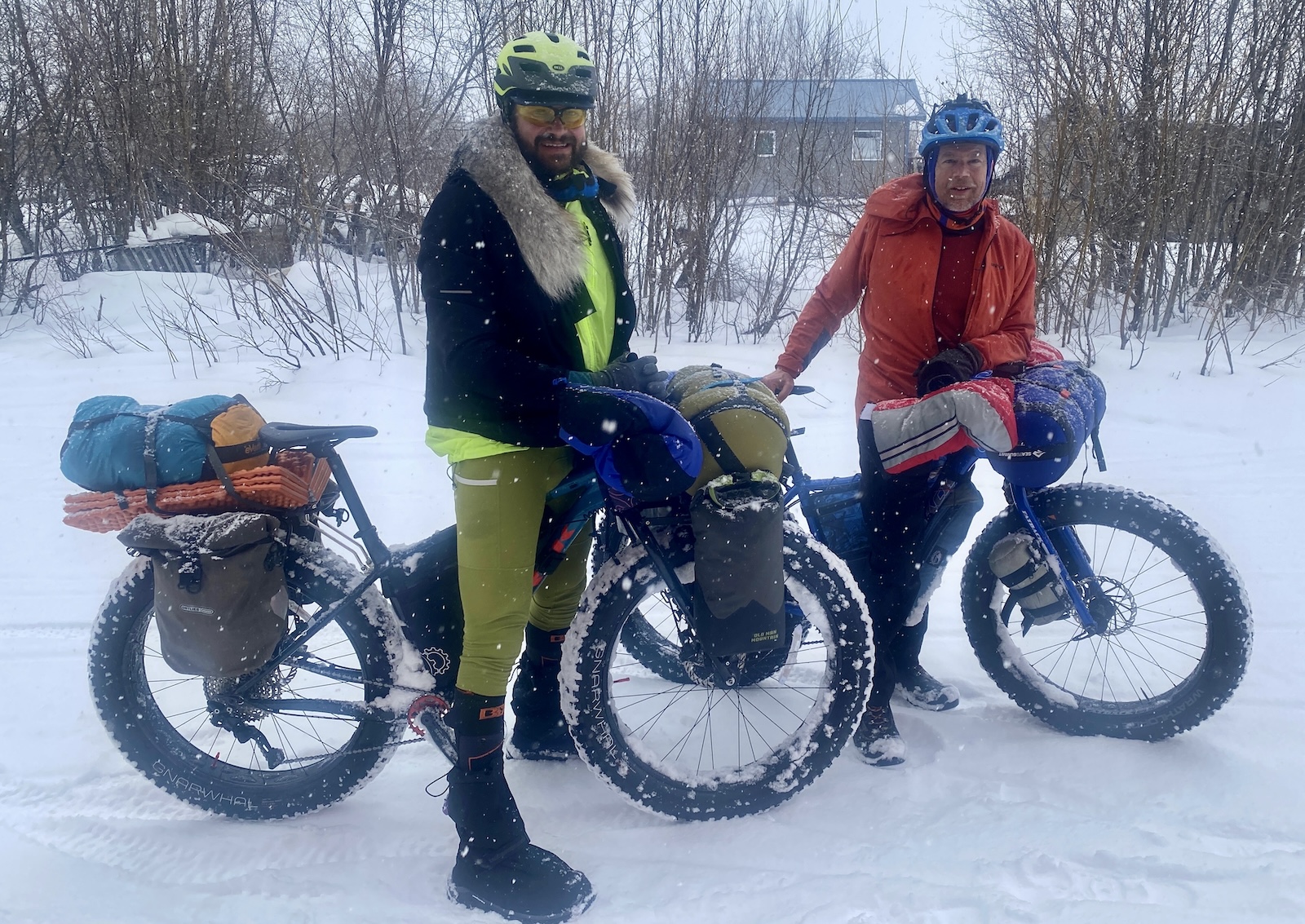 On snowy ground, two helmeted men stand over fat-tired bikes laden with camping gear while snow falls around them. Behind them is a grove of willows, beyond which a few small buildings are visible.