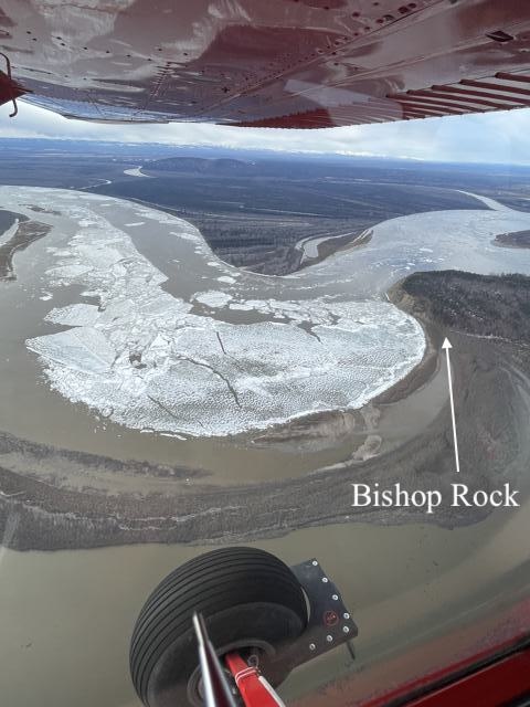 Ice covers the center of a muddy river as it bends sharply and passes by a bluff on the right. The underside of an aircraft wing is visible in the top of the photograph and an aircraft landing wheel is visible in the bottom. 