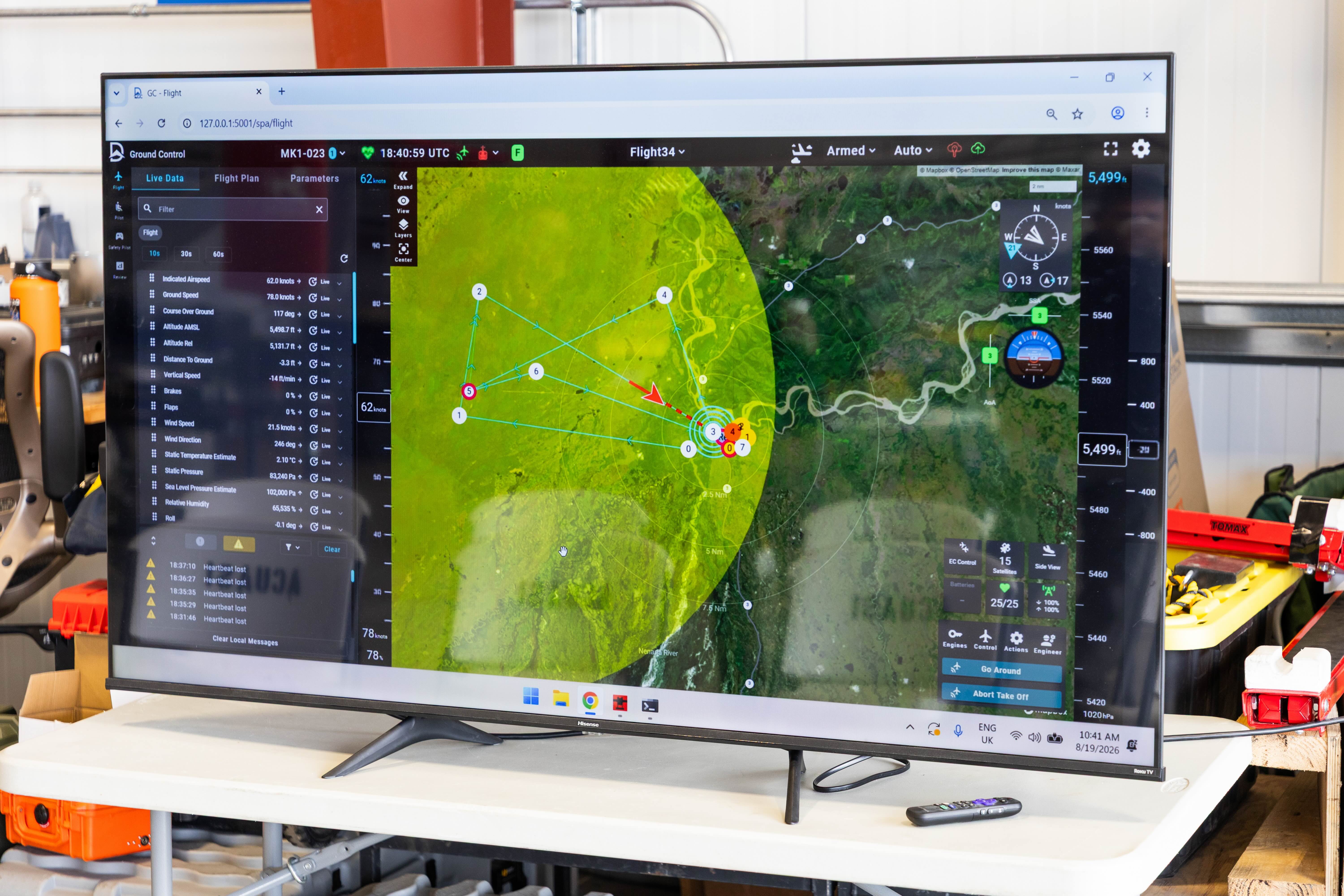 The flight of ACUASI’s Windracers ULTRA drone carrying chilled human blood samples is tracked on a large screen at the program’s Nenana hangar. The display shows the aircraft’s route, position and flight data over a satellite map, with tools, equipment and storage cases visible around the hangar workspace.