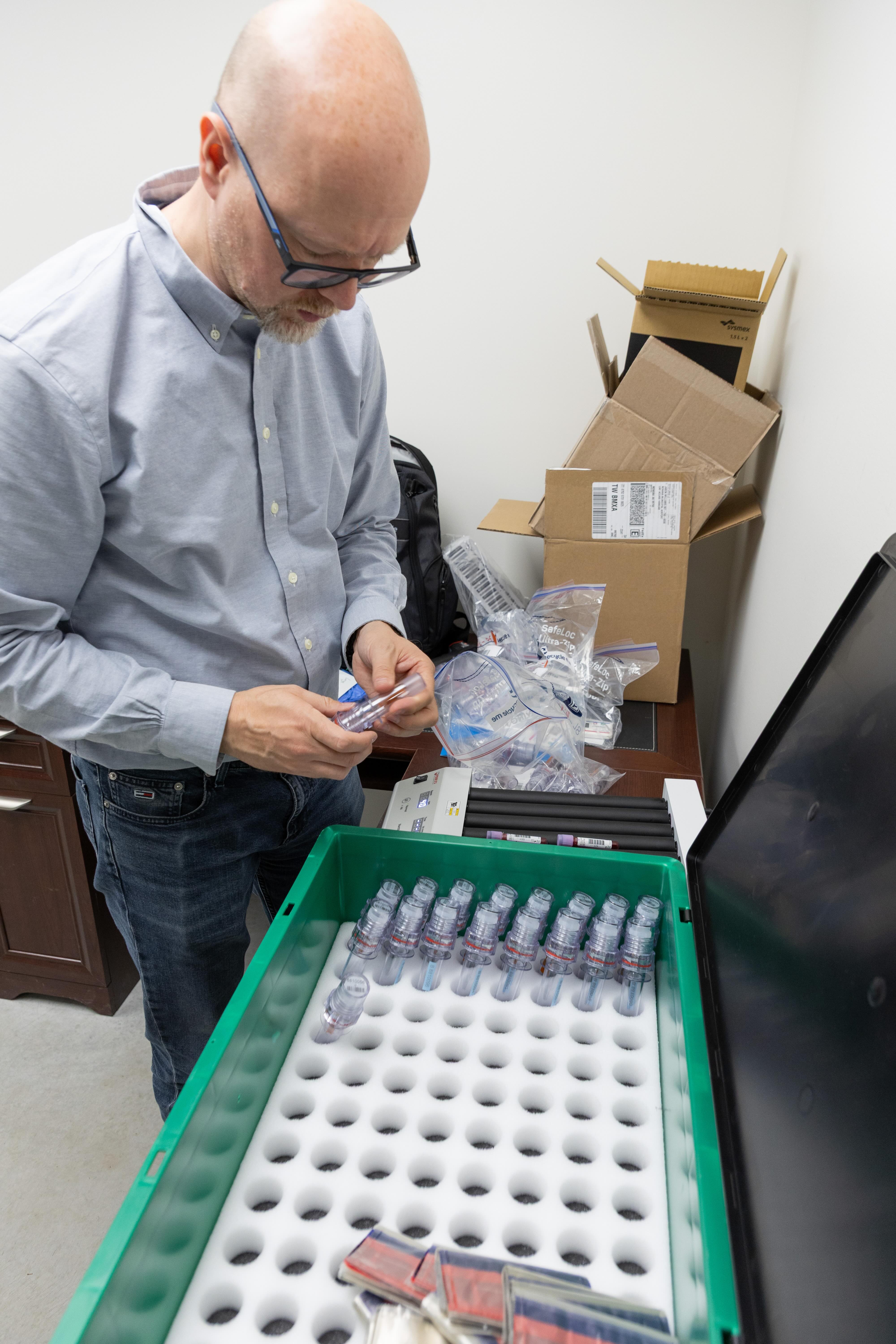 Dr. Sven Voss of the Institute of Doping Analysis and Sports Biochemistry in Germany unpacks blood samples after the 1,000-kilometer flight. He stands beside a green insulated container holding rows of sample tubes in foam inserts, with packing materials, plastic bags and cardboard shipping boxes spread across a nearby desk.