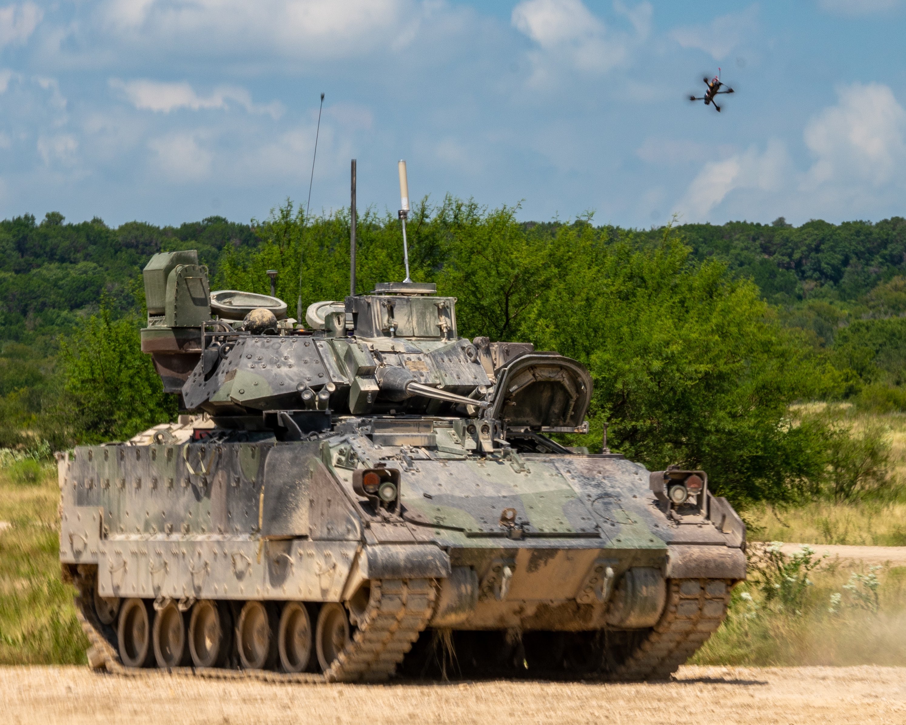 A small quadcopter unmanned aircraft system hovers above an M2 Bradley Infantry Fighting Vehicle on a dirt training range at Fort Hood, Texas, with trees in the background. The scene, captured Aug. 27, 2025, during Operation Return of the Condor, shows the drone positioned overhead as part of testing for drone detection and counter–small UAS tactics.