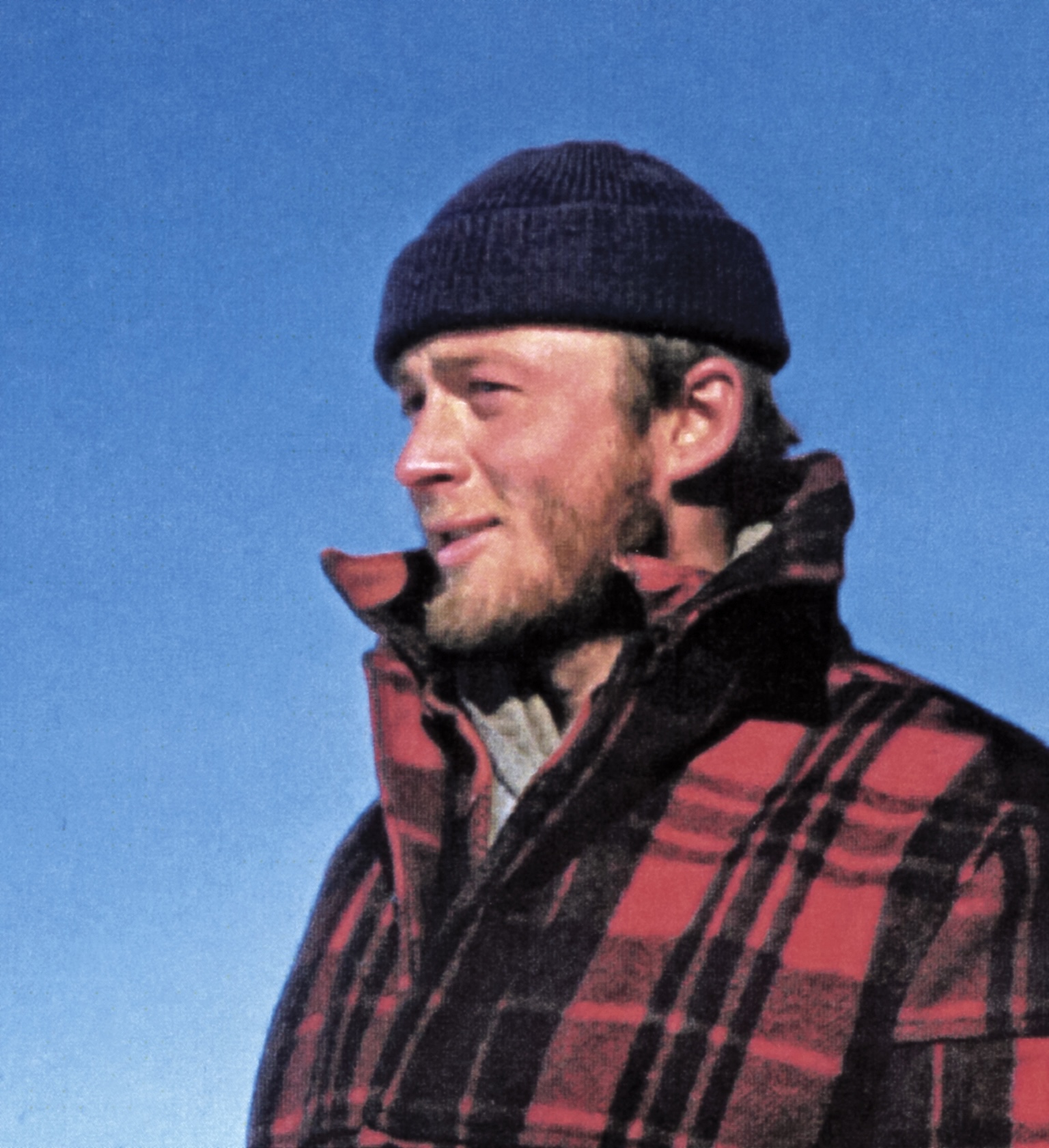 A man with a blond beard stands against a blue sky background while wearing a heavy red-and-black checkered wool coat and a knit cap.