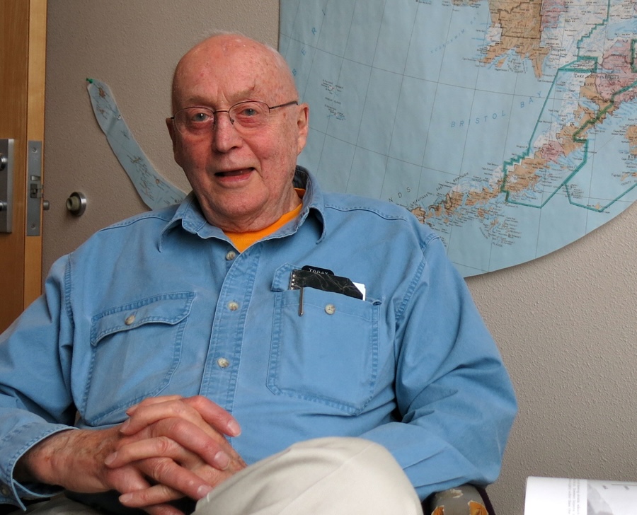 A bald older man wearing glasses and a blue shirt sits in front of a wall with a map of Alaska attached to it.
