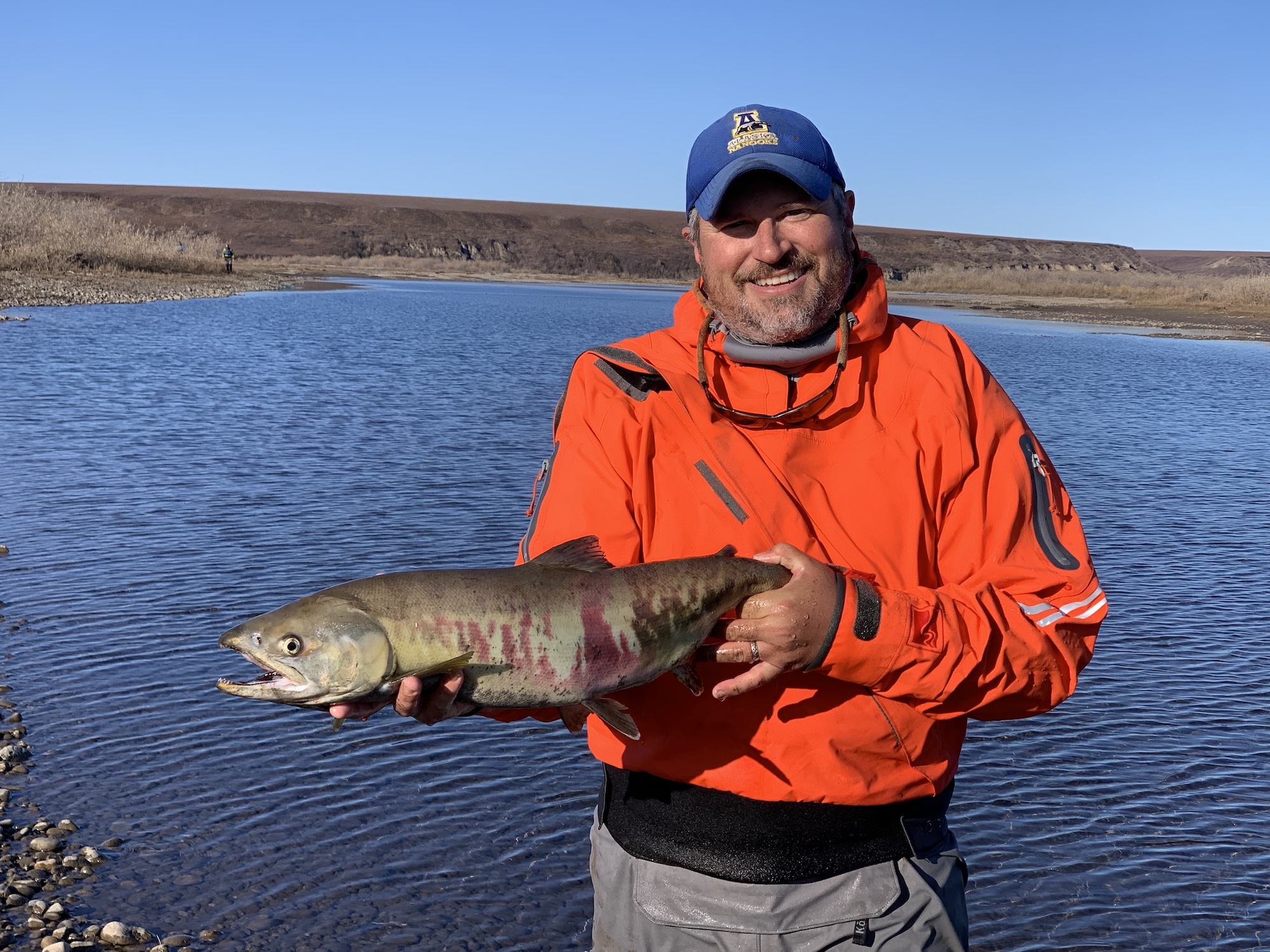 A man in an orange drysuit holds a fish while standing in front of a water body surrounded by a brown tundra landscape.