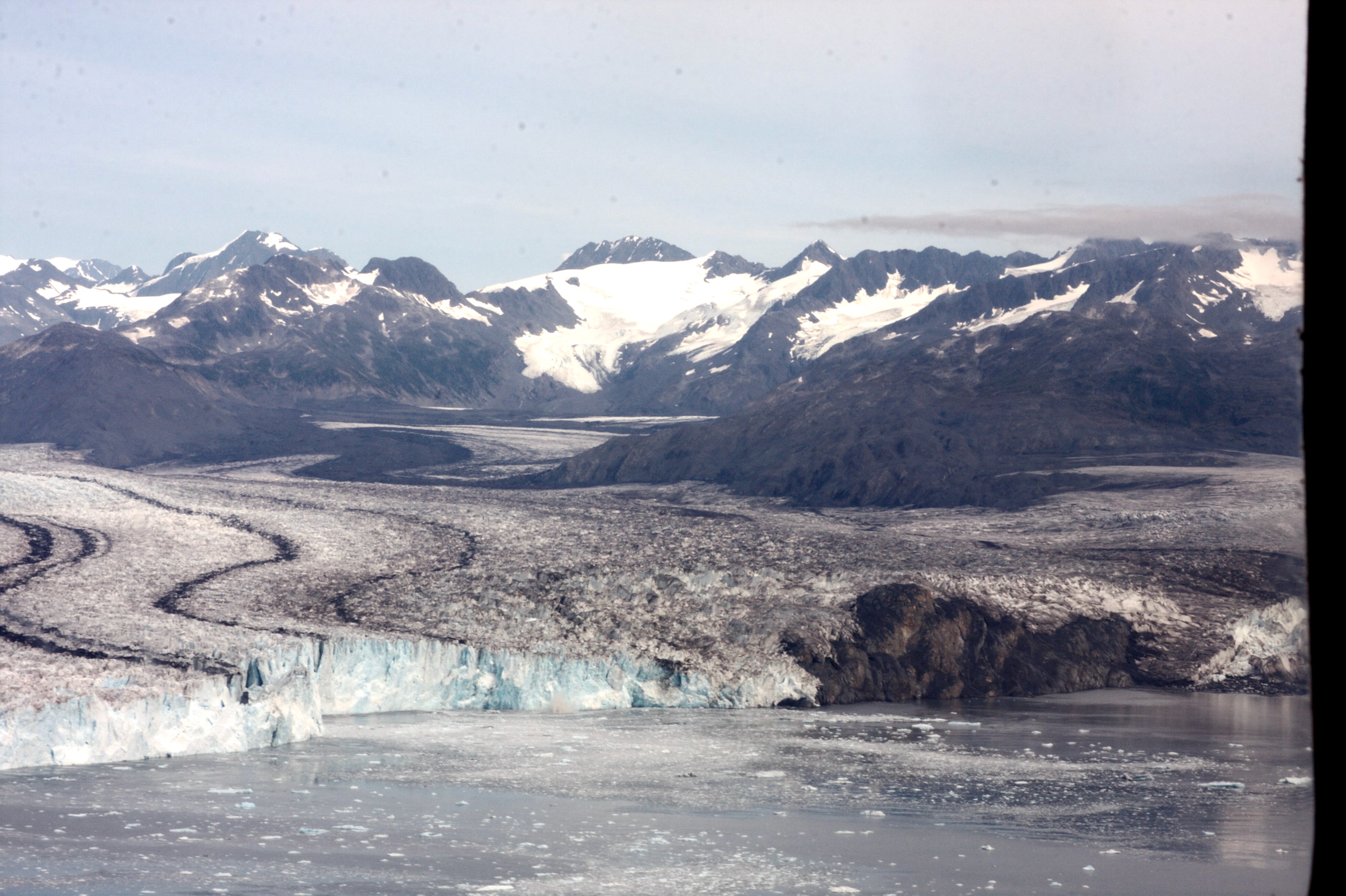 A wide view of Columbia Glacier in Alaska shows a broad river of ice flowing between dark, rocky mountains, with snow-covered peaks in the distance and floating ice in the water at the glacier’s front. Columbia Glacier, shown here in 2016, is about 20 miles west of Valdez and has been retreating since the early 1980s after roughly 200 years of stability.
