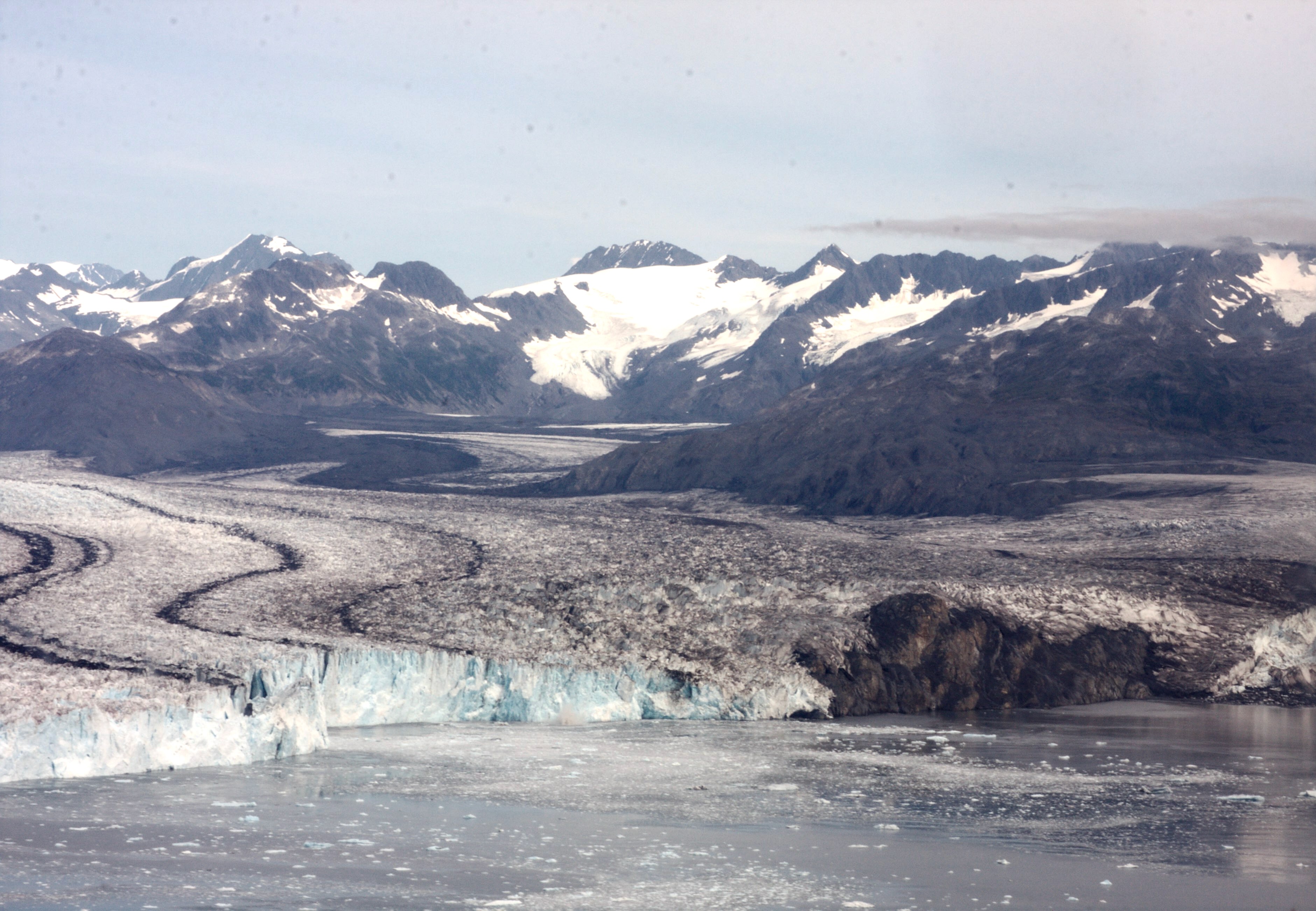 A wide view of Columbia Glacier in Alaska shows a broad river of ice flowing between dark, rocky mountains, with snow-covered peaks in the distance and floating ice in the water at the glacier’s front. Columbia Glacier, shown here in 2016, has been retreating since the early 1980s after roughly 200 years of stability.