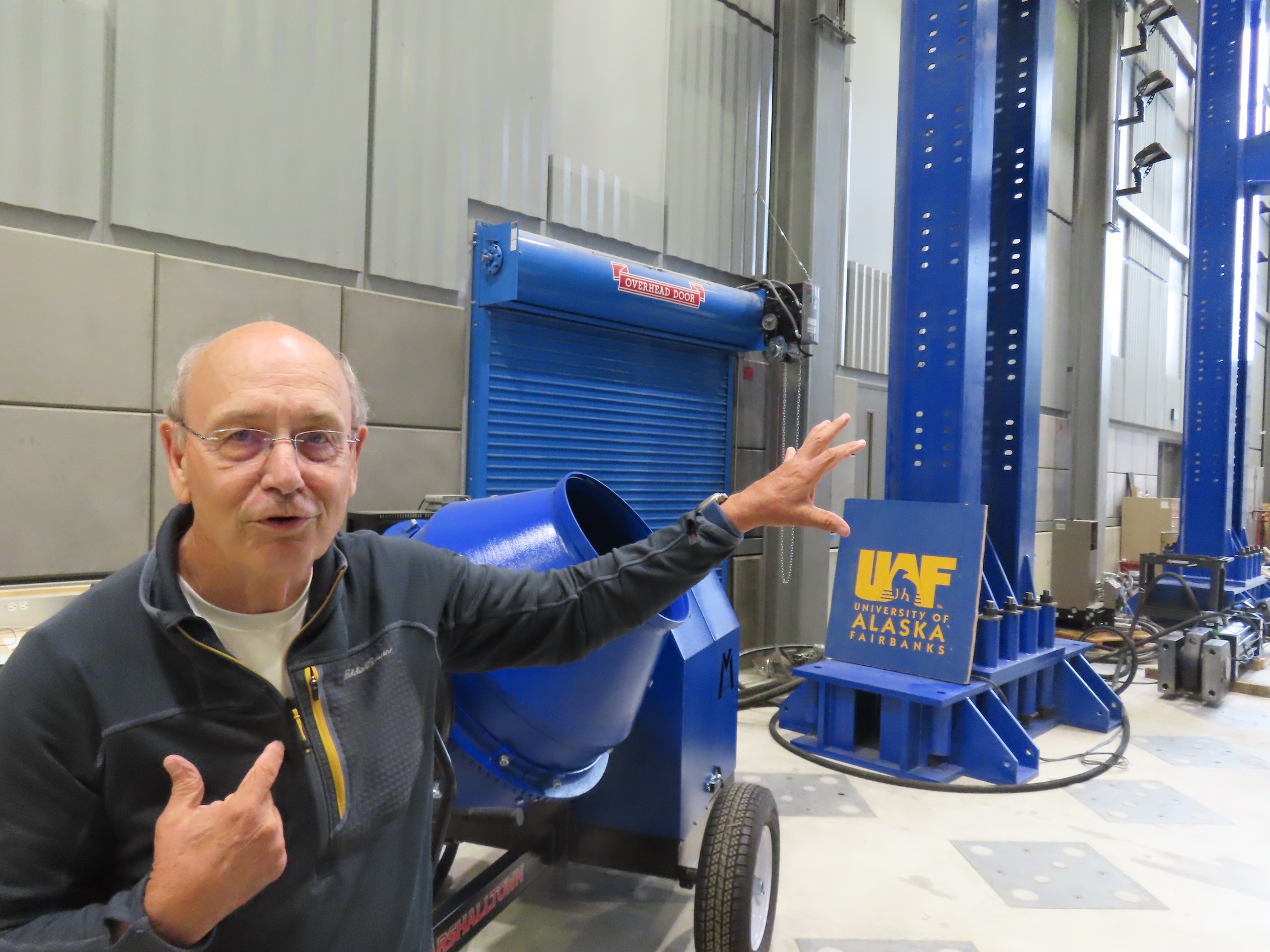 A man in a blue sweater gestures toward large steel beams, painted blue, in a high-ceilinged construction materials testing laboratory.