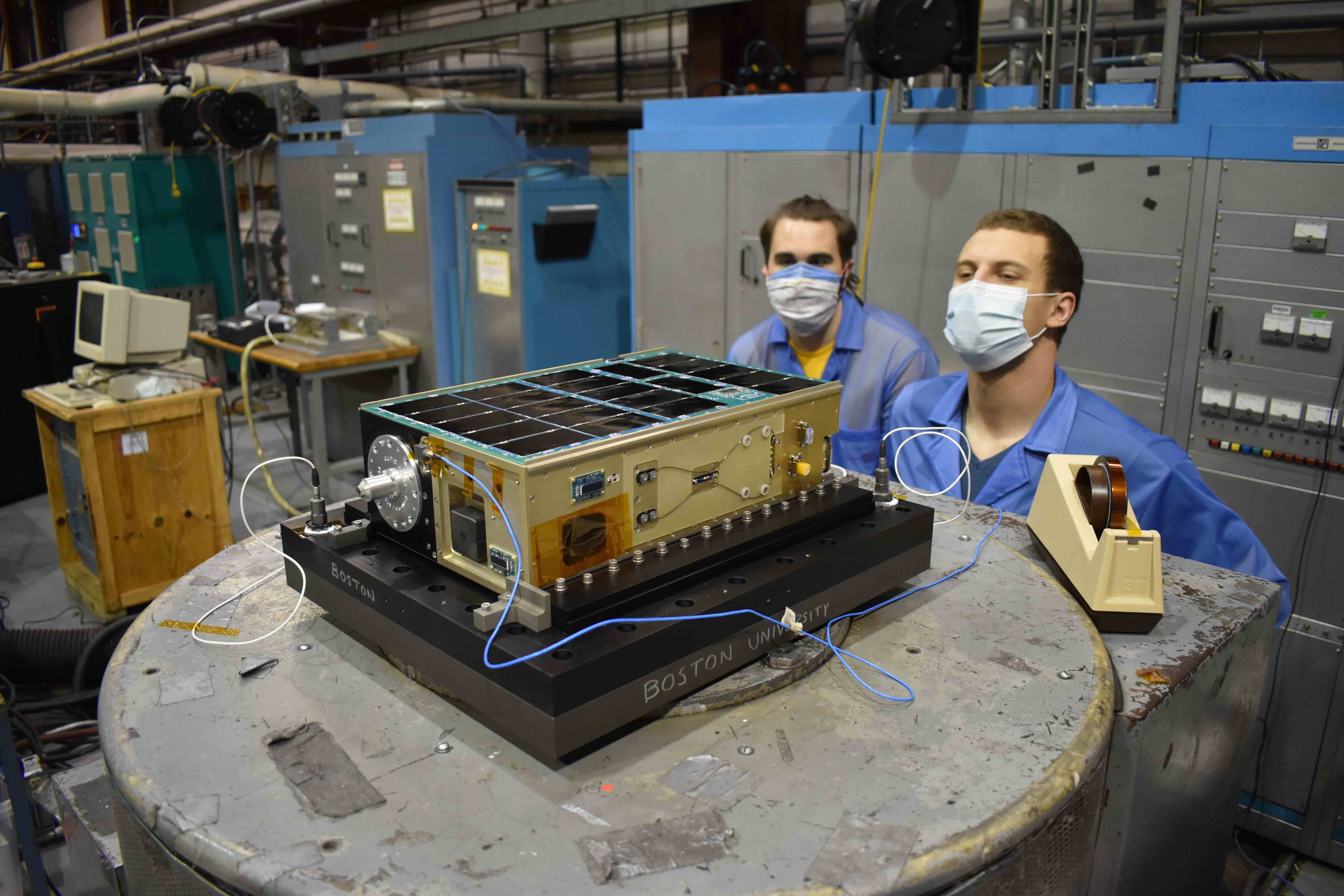 Two men in face masks work on a small satellite, called a cubesat, on a table in a workshop.