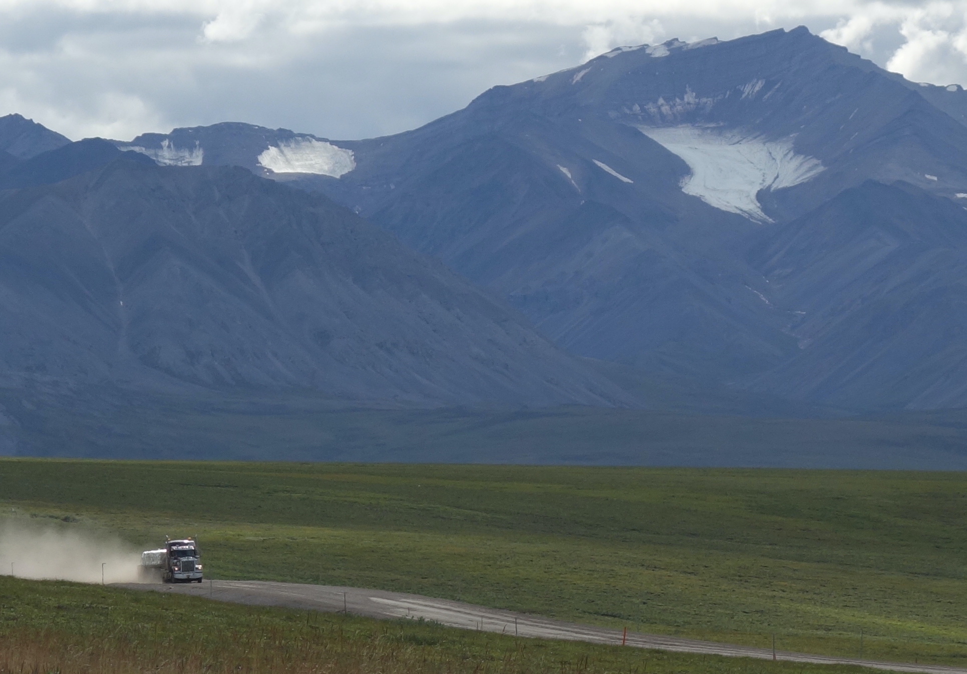 A tractor trailer drives through green tundra with mountains bearing a few small glaciers in the background.