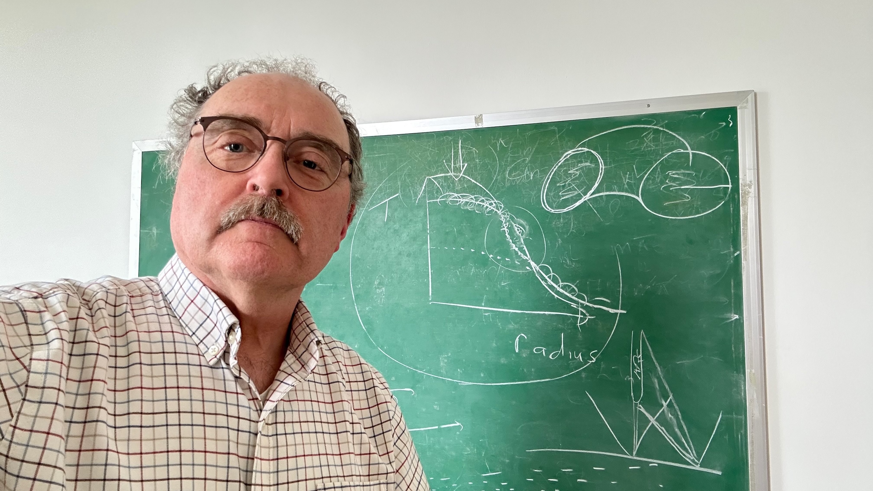 A man with glasses and a mustache stands in front of a green chalkboard with scrawled diagrams.