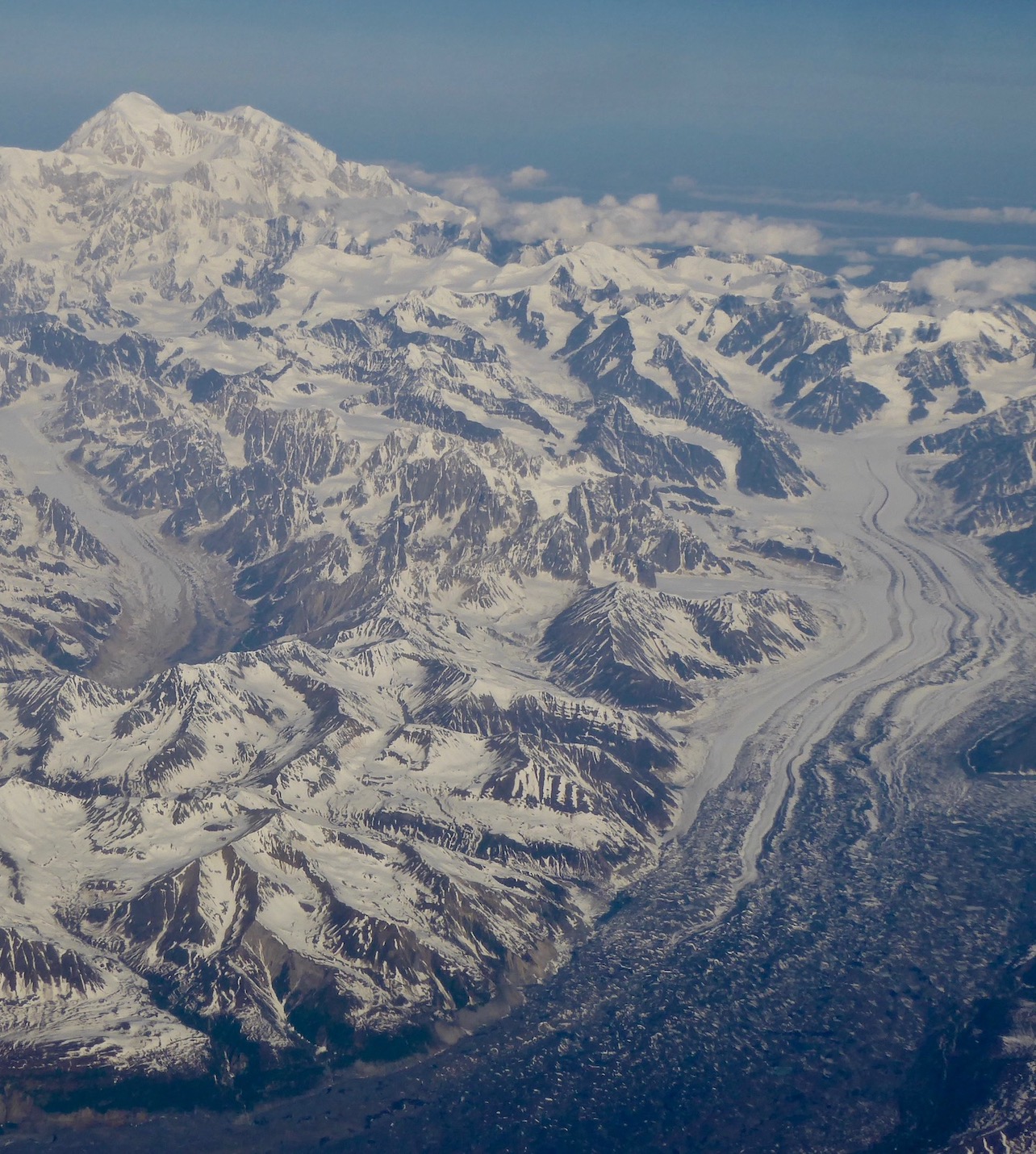 A large snow-covered mountain rises above lower peaks and glaciers in the foreground in an aerial view.