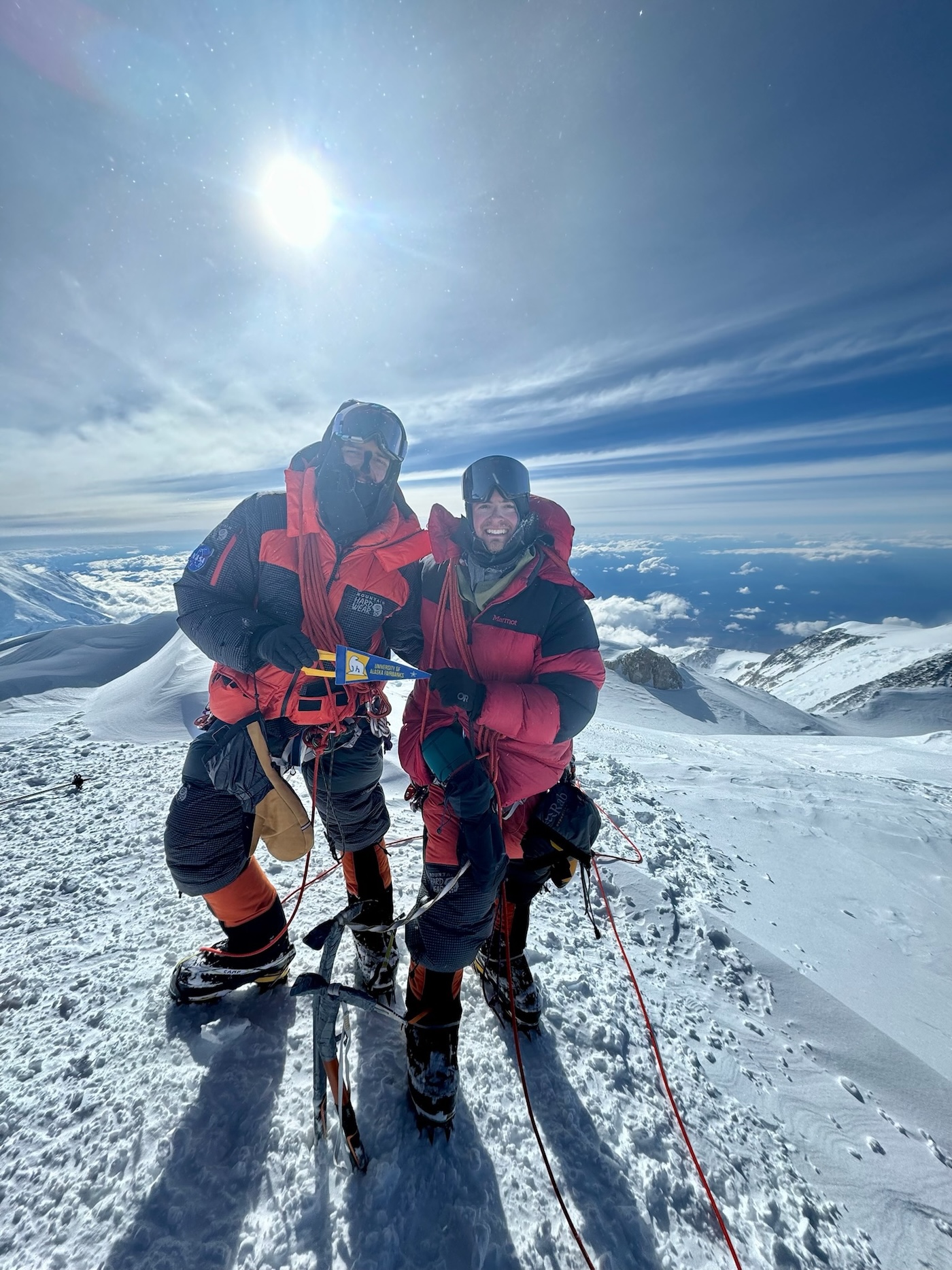 Two men in winter gear stand together on a snowy mountain top with the sun shining behind them.