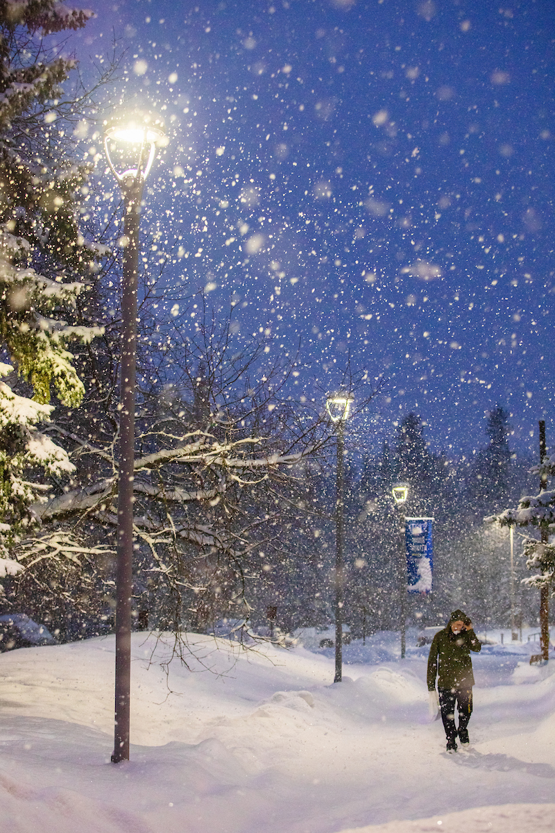 A person walks through snowfall under lighted streetlamps on the UAF campus.