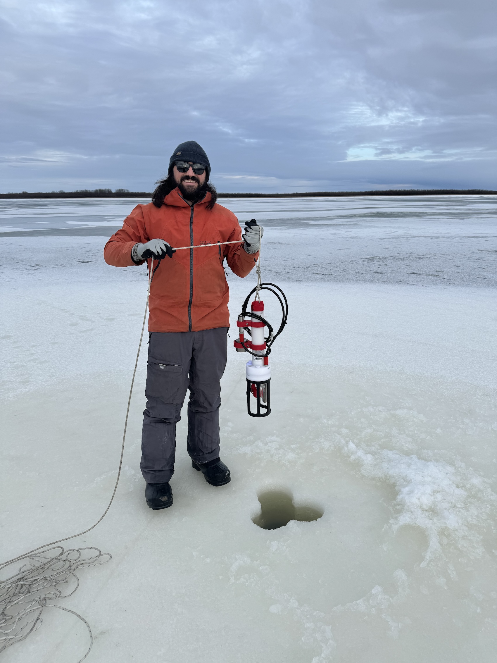 A man stands near a hole in a sheet of river ice while holding an instrument.