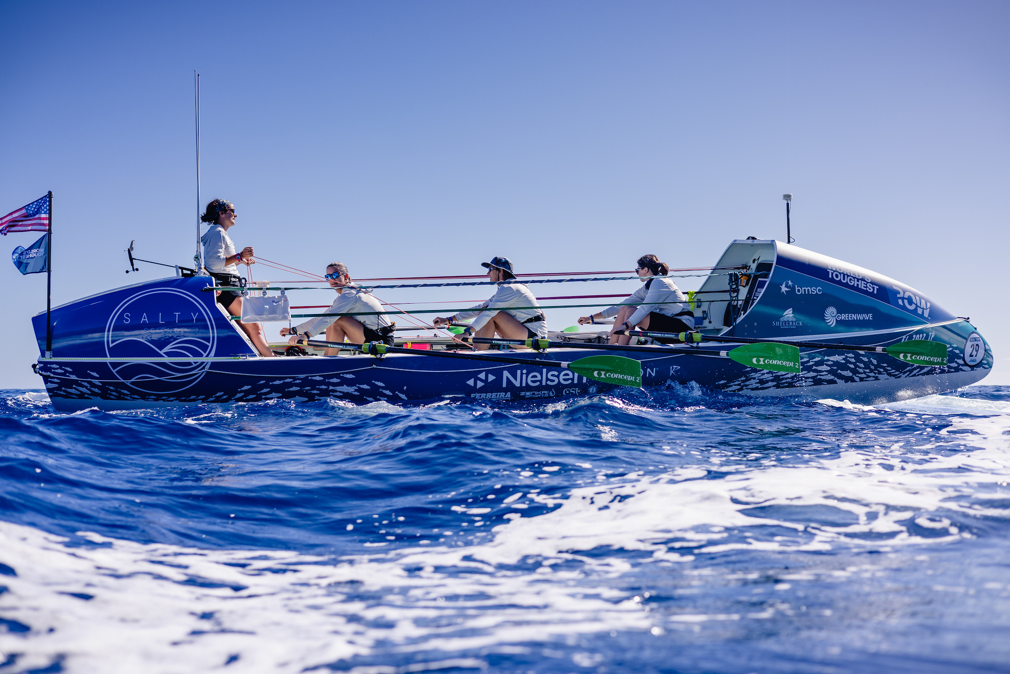 Four women, three rowing and one steering, propel a boat across ocean waves.