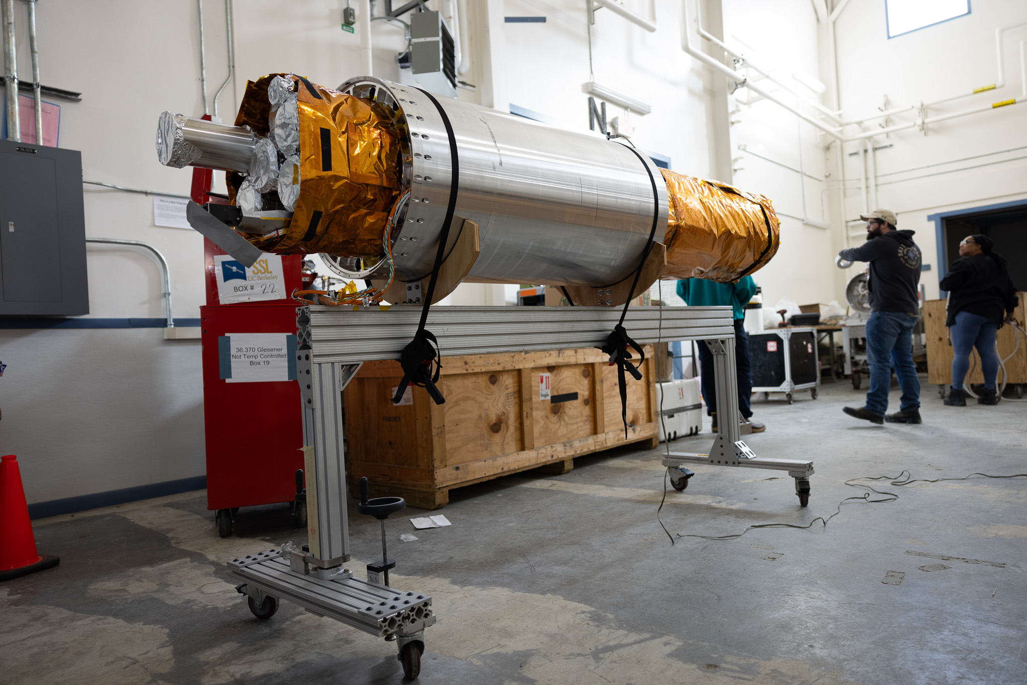 The payload section of the FOXSI-5 mission rests on a wheeled support frame inside the payload assembly building at Poker Flat Research Range on April 13, 2026. The cylindrical instrument, wrapped partly in gold thermal insulation, is surrounded by equipment crates and technicians preparing it for flight. The NASA sounding rocket carrying the payload is scheduled to launch in early May.