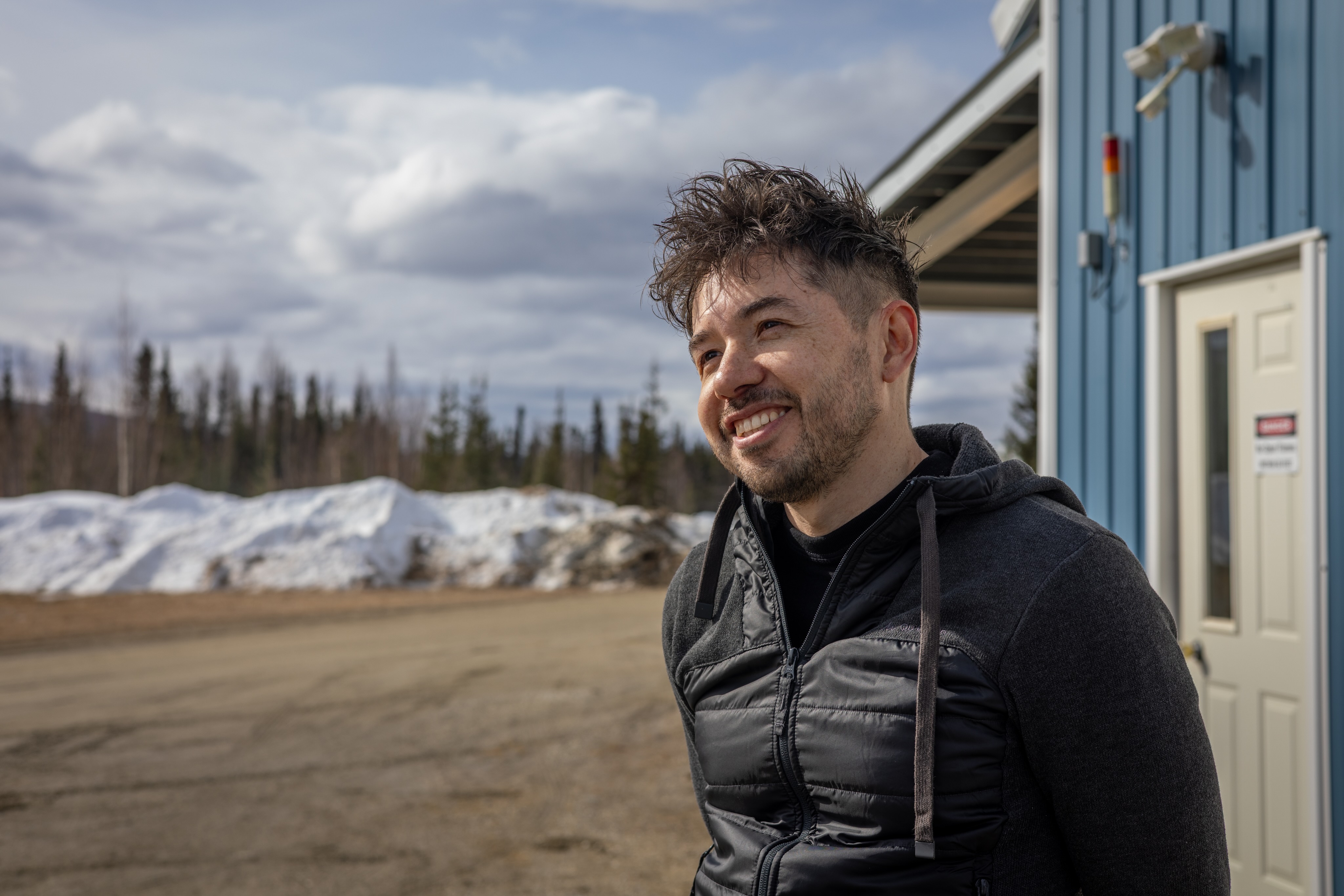 Juan Camilo Buitrago-Casas of the University of California, Berkeley’s Space Sciences Laboratory stands outside the payload assembly building at Poker Flat Research Range on April 13, 2026, speaking about the FOXSI-5 mission he leads. He smiles while facing slightly to the side, with snowbanks, trees and a partly cloudy sky in the background.