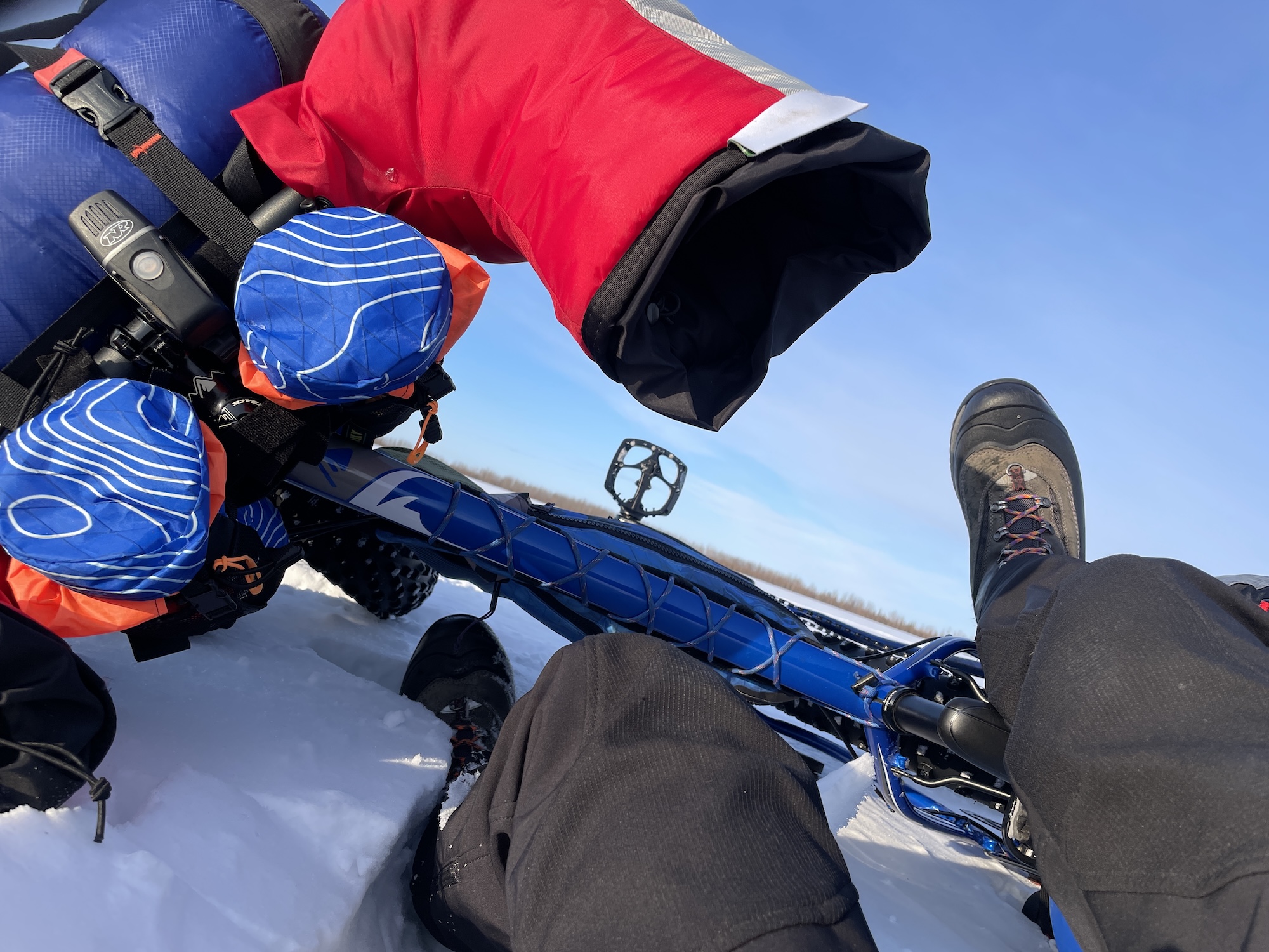 A person's legs and feet stick into the air around a bicycle frame lying on its side in the snow. Large overmitts, called pogies, cover the bike handlebars.