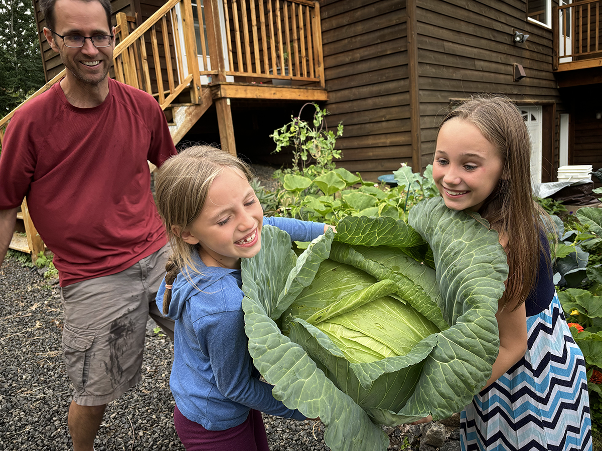 A dad watches as two children hold a giant cabbage. 