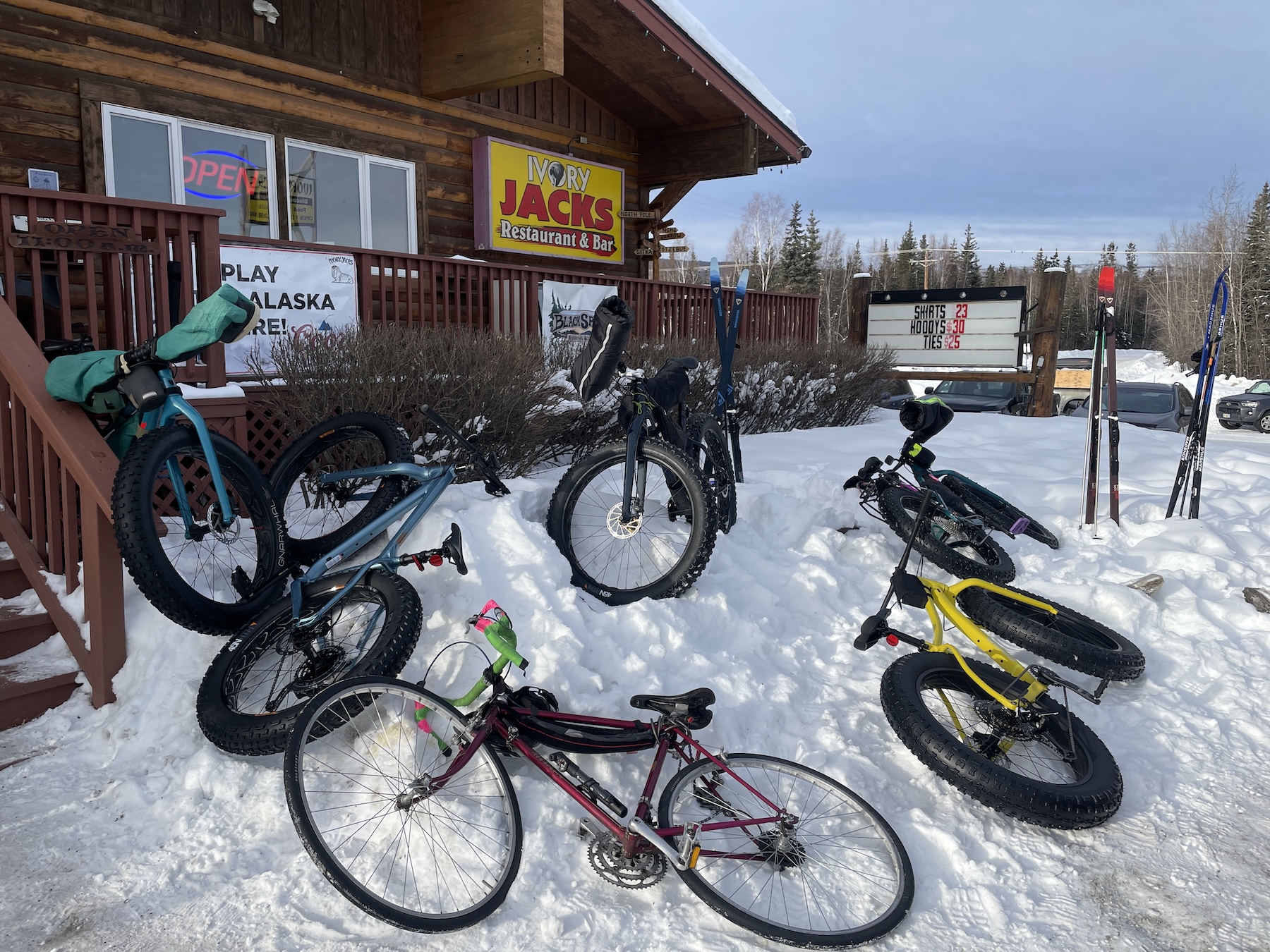 Several bicycles lie on a snowbank next to a building with a raised wooden deck and railing along its perimeter.