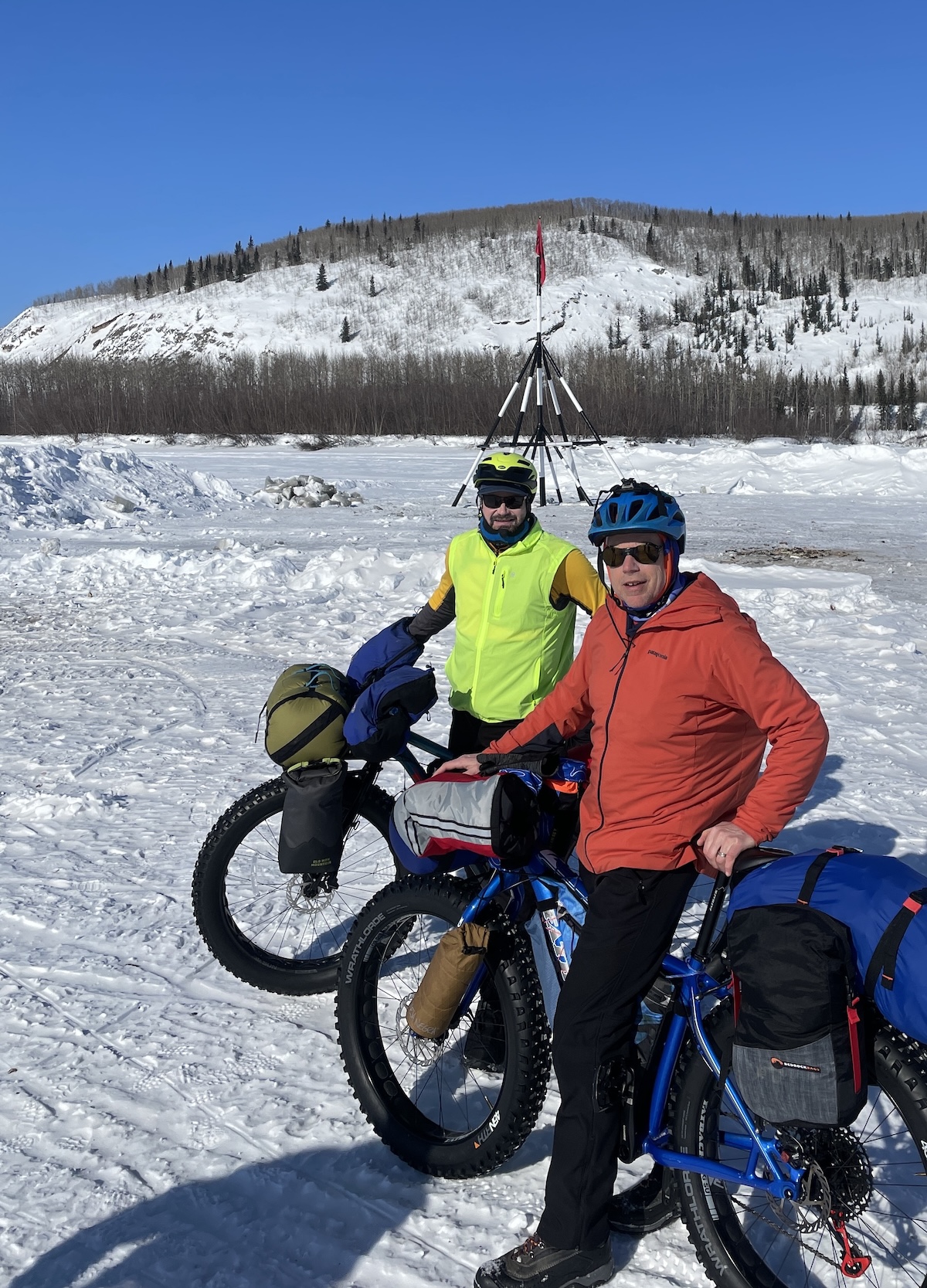 Two men straddle fat-tired bikes strapped with gear bags on an area of packed snow. Behind them, a black and white tripod of poles, topped with a red flag, rests on the surface of a frozen river. Beyond that, a line of cottonwood trees grows above the far riverbank, and, beyond that, a rocky bluff and lightly forested hill rise into a blue sky.