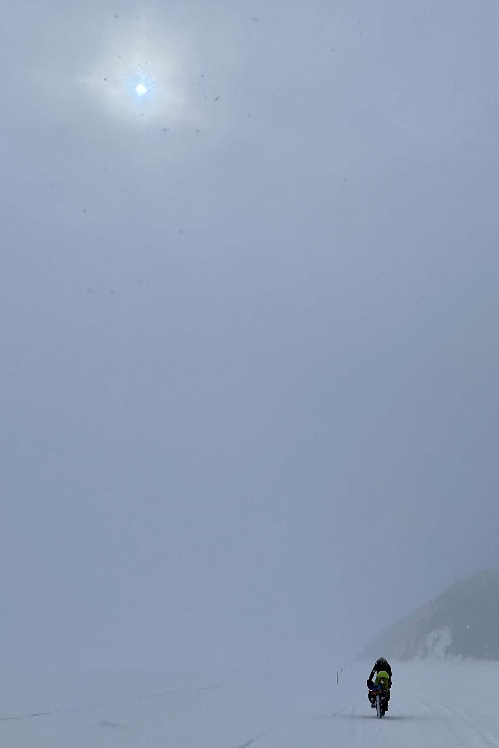 The sun shines through thin clouds and fog above a bicyclist riding on a snowy river. In the background, a bluff emerges from the fog on the right.
