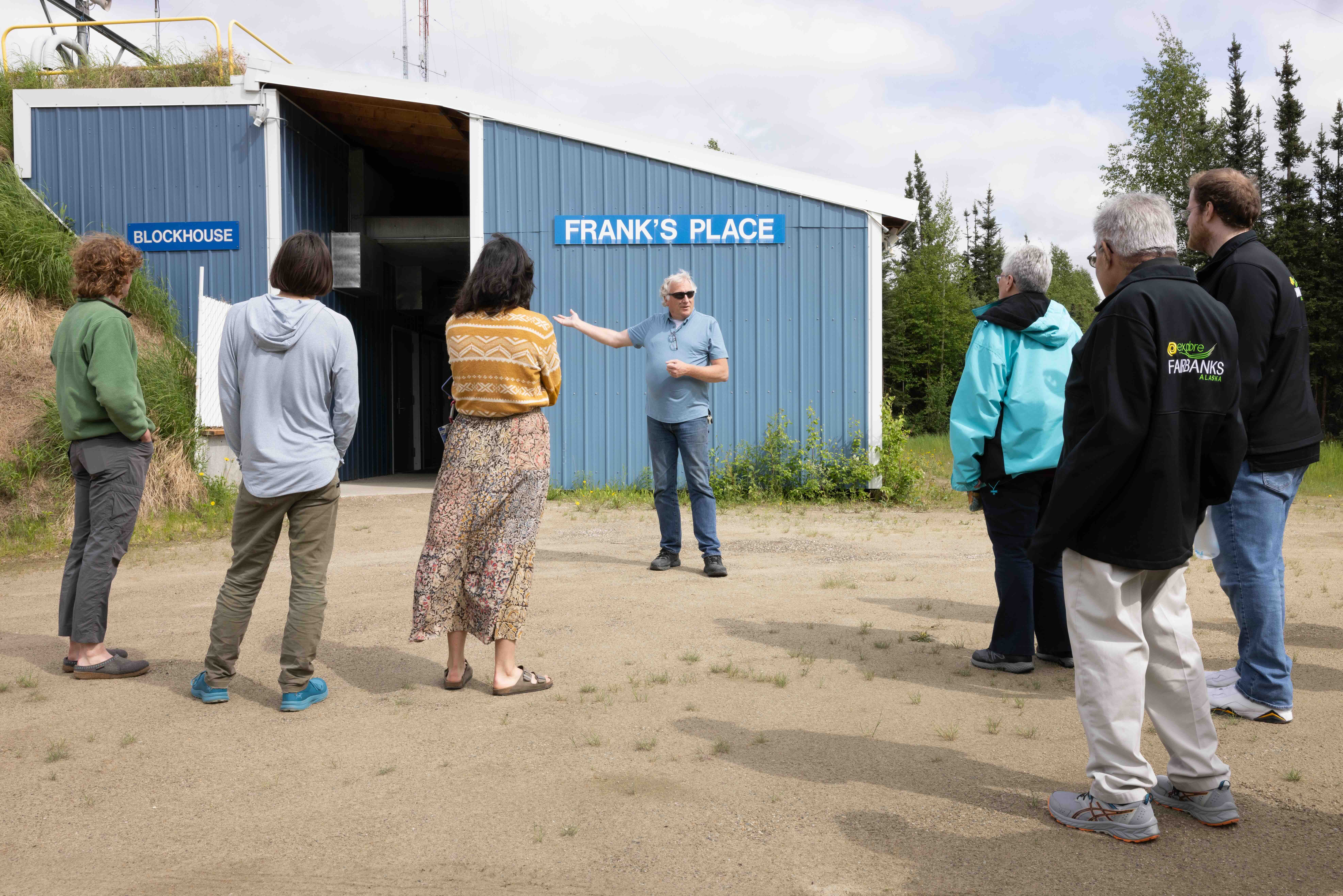 Dave Hill explains the function of the Blockhouse Complex during a public walking tour of Poker Flat Research Range on June 13, 2024. Hill gestures toward the blue Blockhouse Complex building as tour participants stand outside listening on a gravel roadway surrounded by trees and grassy berms.