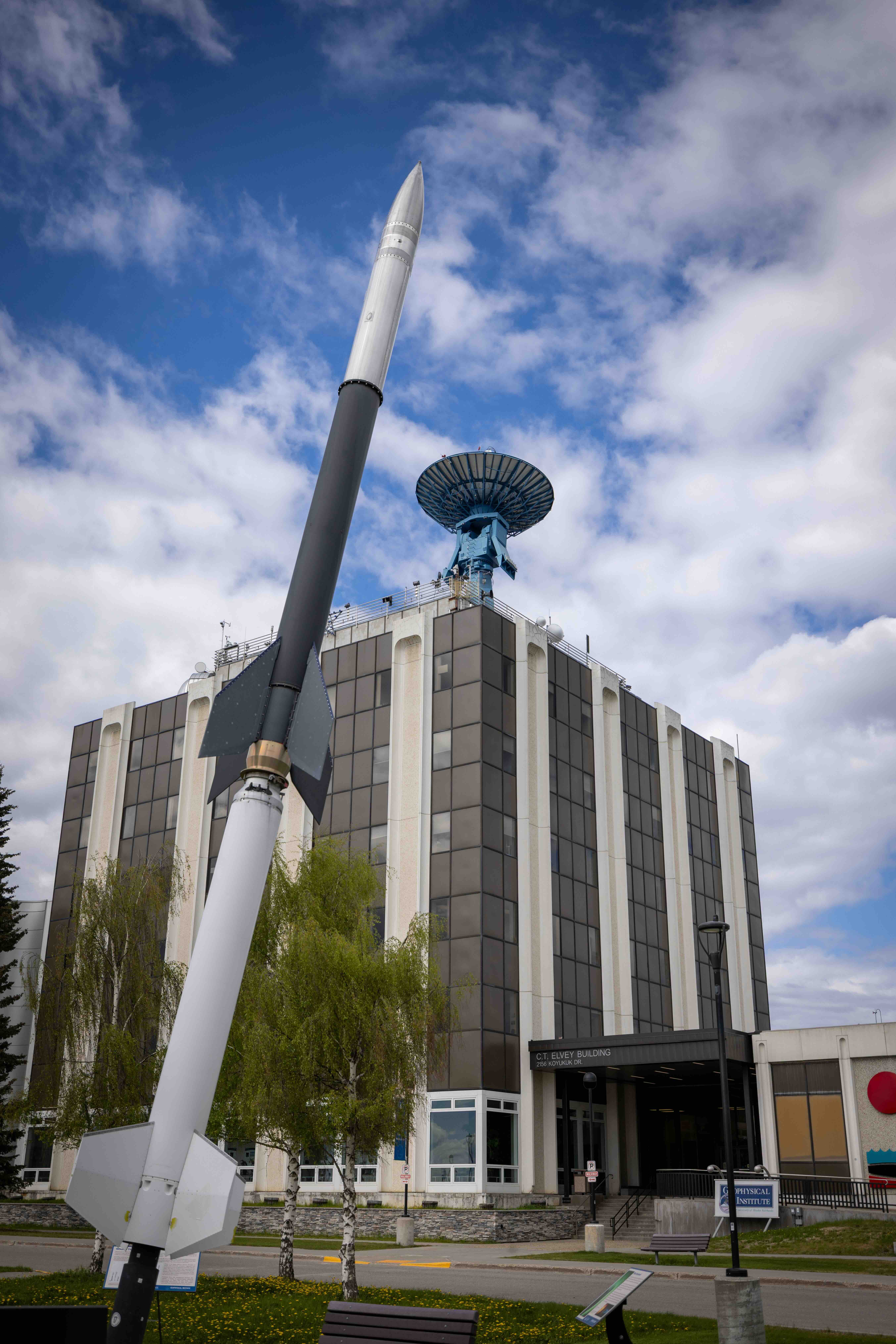 Visitors on the Geophysical Institute summer tour will learn about the large blue satellite antenna atop the Elvey Building and the sounding rocket installation displayed on the lawn outside. The photo shows the rocket model rising in the foreground in front of the University of Alaska Fairbanks Geophysical Institute building under a partly cloudy sky.