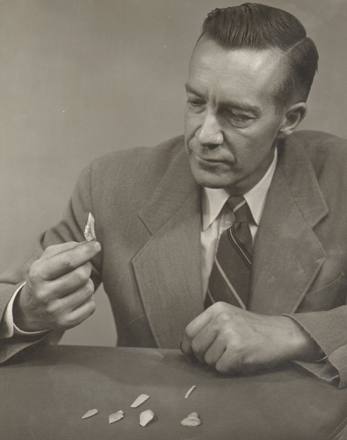 A wearing a suit and tie sits at a table while examining a shaped stone point. Other stone points and tools sit on the table.