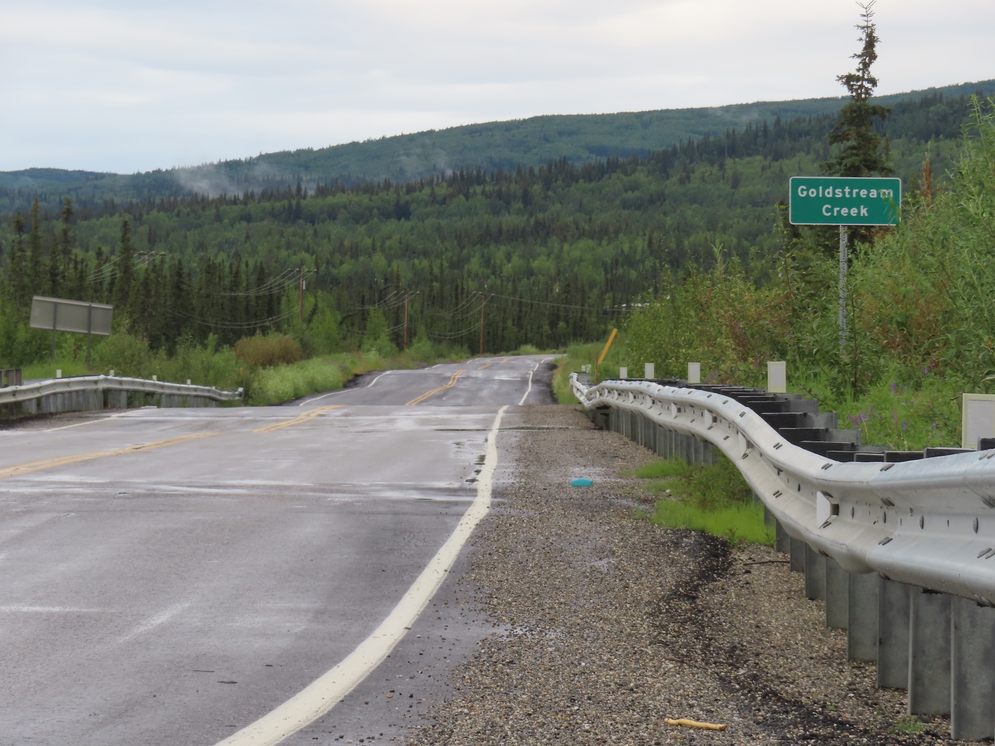 A bumpy road is accompanied by an equally distorted guardrail as it traverses a green forested valley. A green road sign to the right of the road bears the name "Goldstream Creek," indicating the location of the creek and bridge.