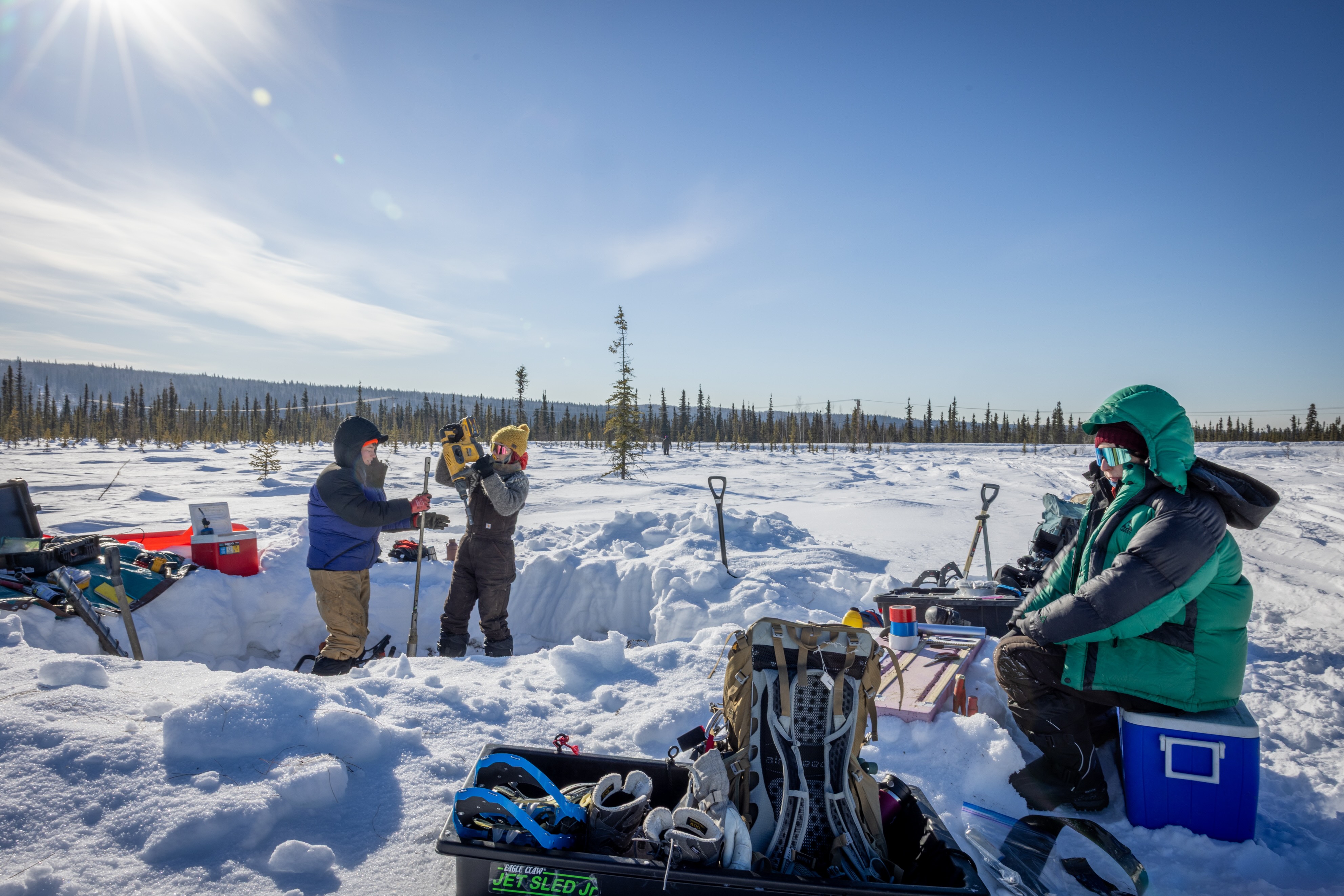 Three bundled researchers work at a snow-covered permafrost coring site in a boreal forest near Fairbanks under a bright, low winter sun. From left, Louise Farquharson of the University of Alaska Fairbanks Geophysical Institute, and Shannon Hibbard and Jennifer Kielhofer, both of the Desert Research Institute, operate coring equipment beside a dug trench, with shovels, packs and field gear spread across the snow in the Goldstream Creek area on April 13, 2026.