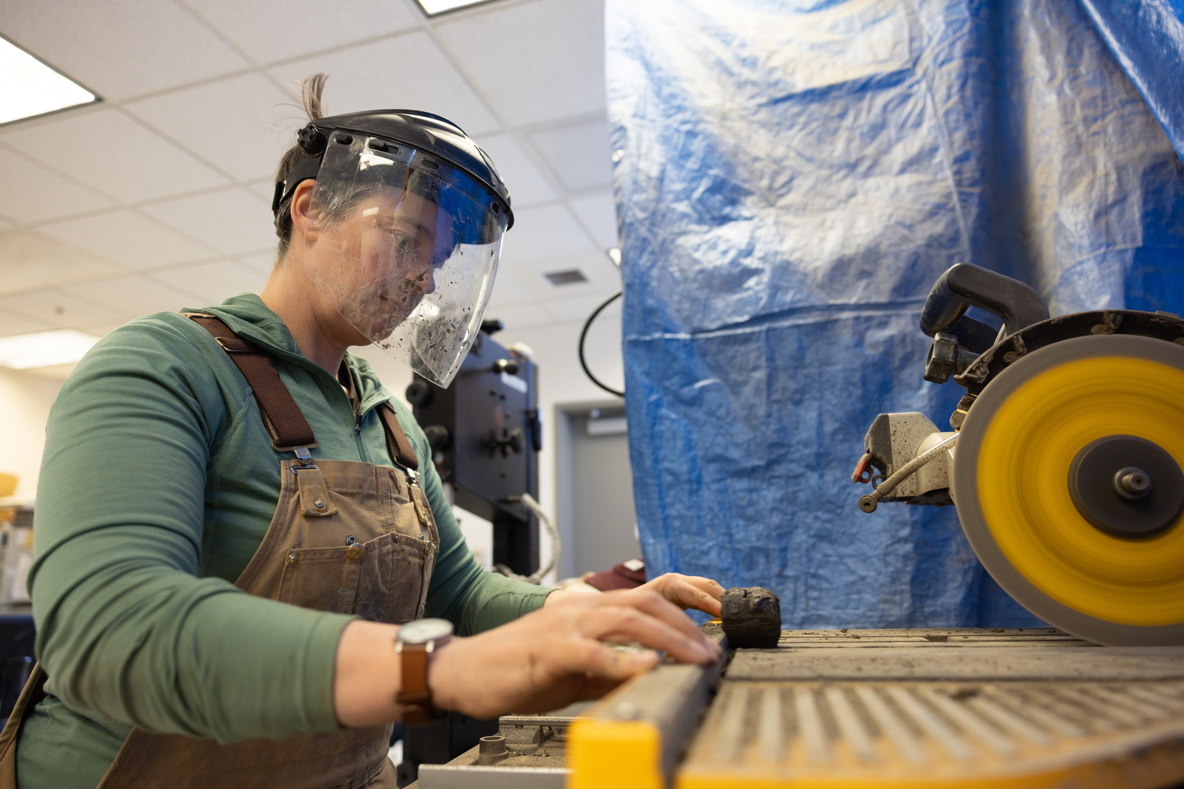 UAF research assistant professor Louise Farquharson wears a face shield and apron while cutting a dark core sample on a spinning saw in a laboratory at the UAF International Arctic Research Center. The close-up shows her guiding the sample carefully along the blade, with equipment and a blue protective curtain in the background.