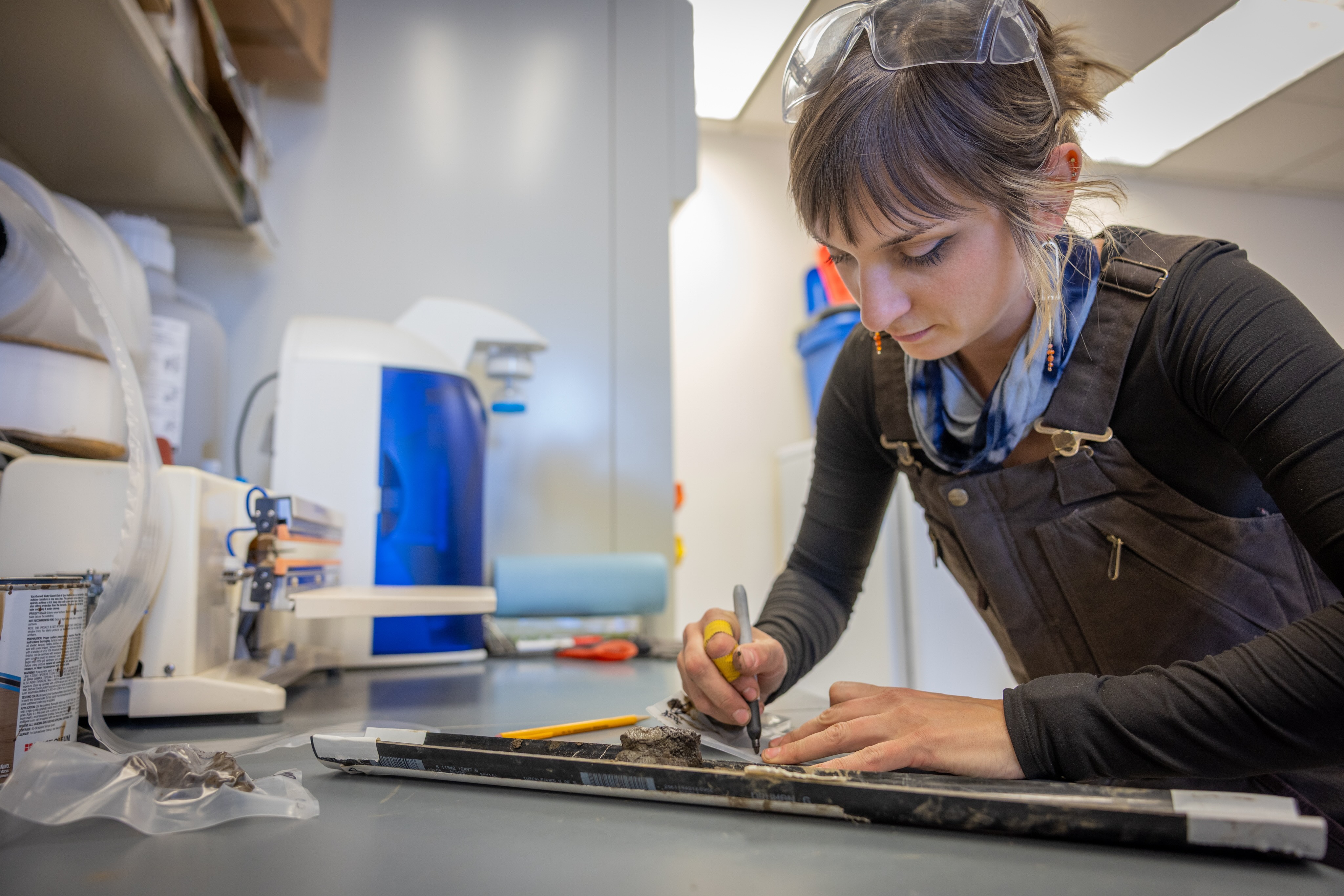 Assistant research professor Shannon Hibbard of the Desert Research Institute leans over a lab bench, carefully labeling and packaging a split core sample for storage. She wears safety glasses and work overalls, using a marker to note details on the sample while lab equipment and containers sit nearby.