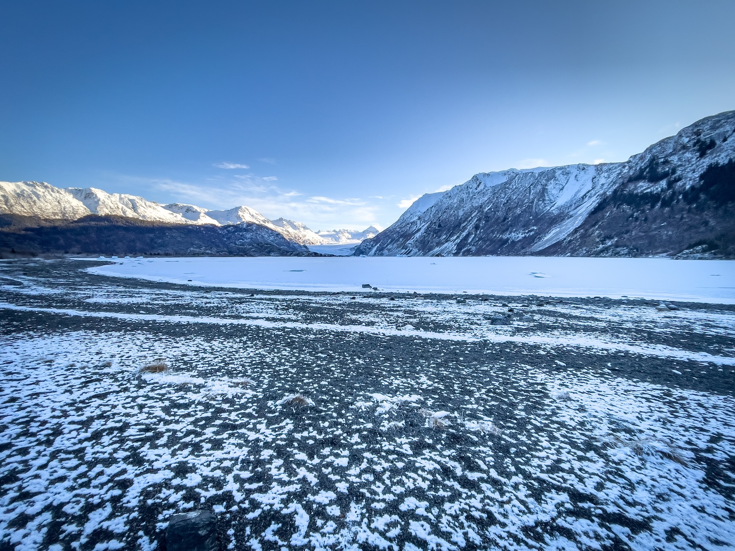 A thin layer of snow covers a sandy beach and a frozen lake, with sunlit mountains in the background.