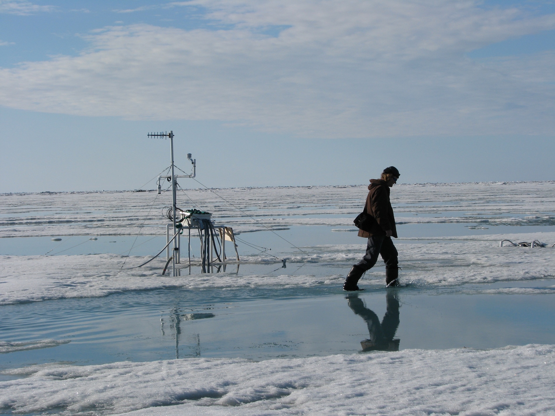 On a sunny day, a man walks through a puddle on an expanse of sea ice. An array of instruments sits on the ice behind him.