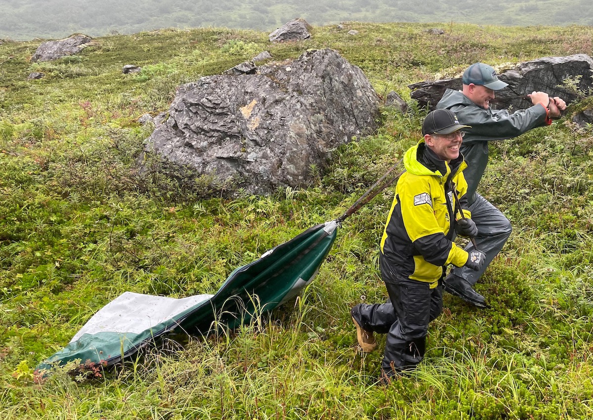 Two men drag a deflated boat pontoon across green alpine tundra.
