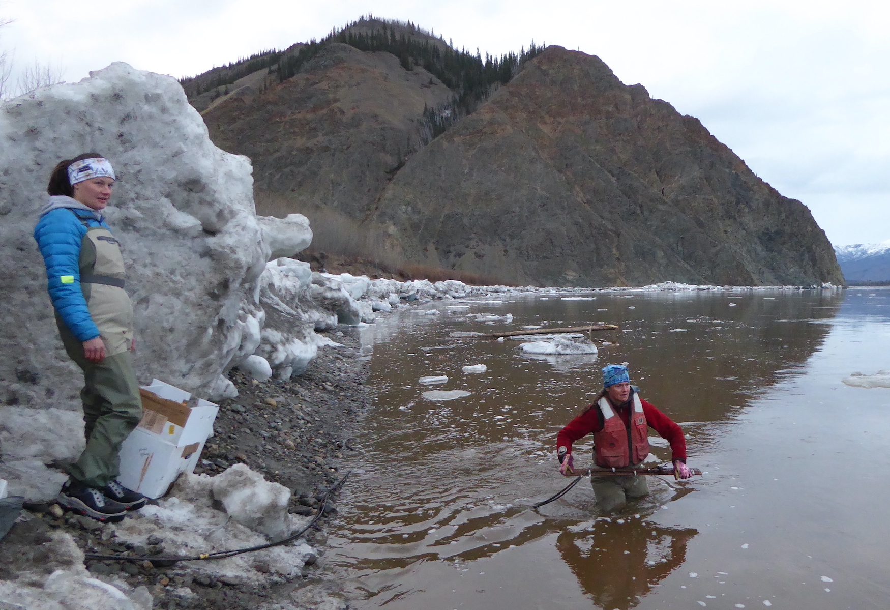 A woman stands in a river with ice chunks on its banks while another woman watches from the bank.