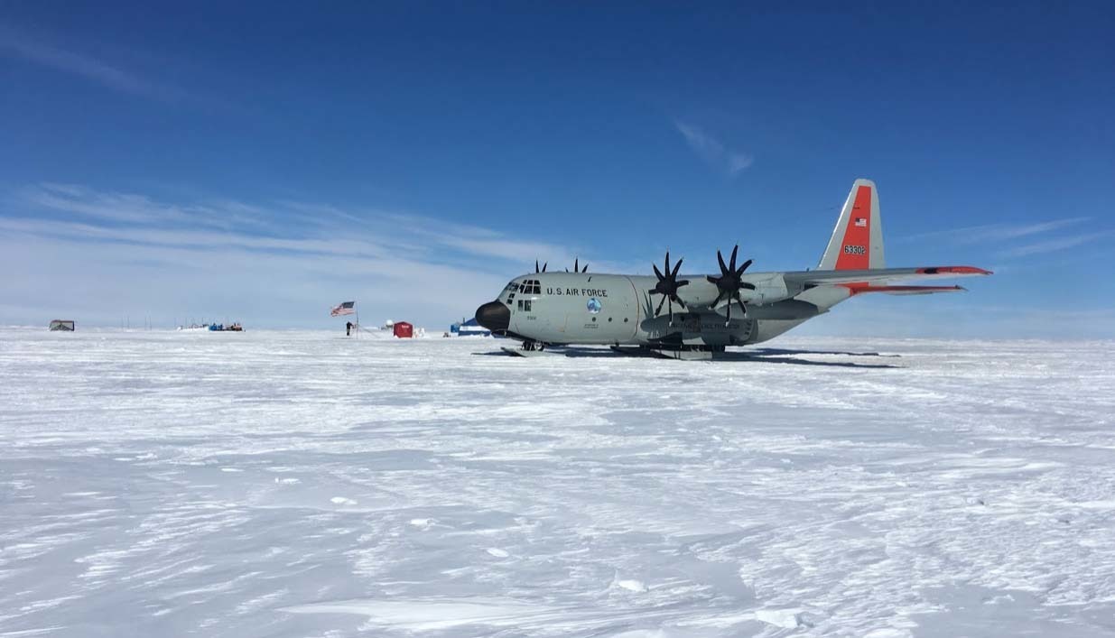 A New York Air National Guard LC-130 Skibird cargo aircraft sits on a snow runway atop the Greenland Ice Cap under a clear blue sky. The ski-equipped aircraft, operated by the 109th Airlift Wing, is parked on the windswept ice with a small field camp, flags and support structures visible in the distance. The photograph was taken during Exercise Polar Reach in 2019, a training mission that prepares aircrews for operations in polar environments.