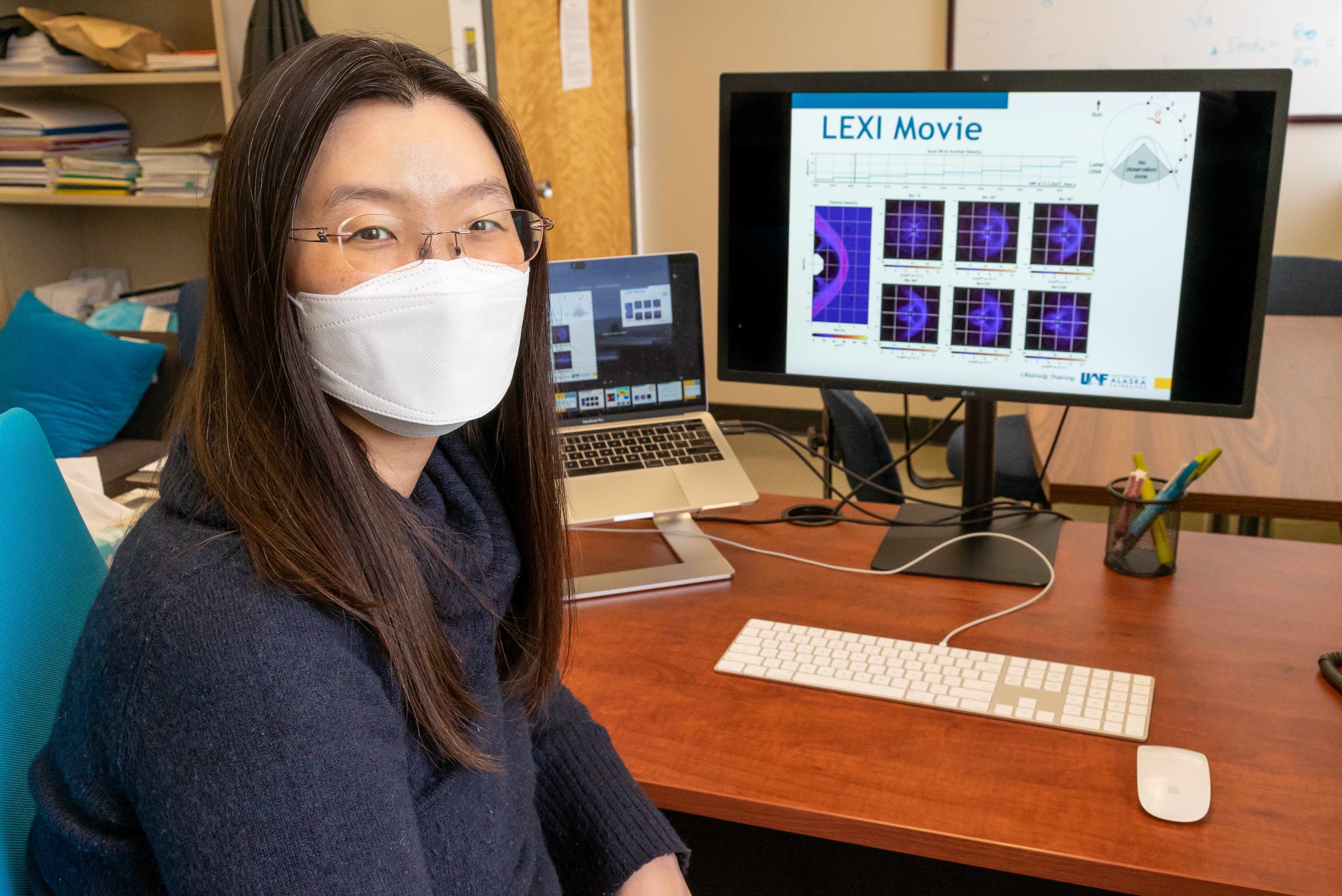 A woman with dark hair and glasses, wearing a face mask, sits at a desk with a computer on it.