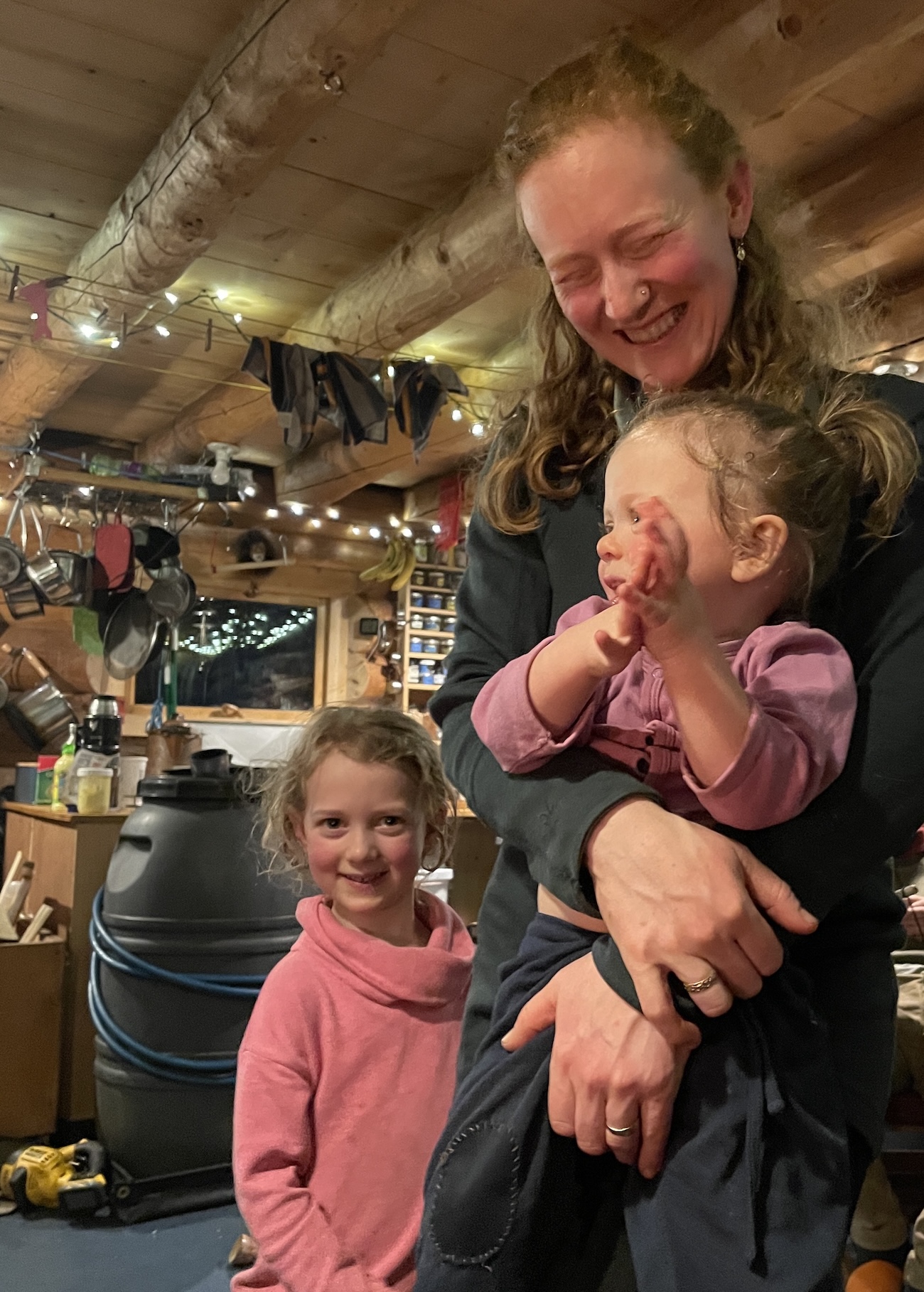 In a log cabin, a woman laughs while holding a toddler. A young girl stands next to her smiling and looking at the camera. In the background, a black plastic water barrel wrapped in a blue hose sits next to a kitchen counter. Peeled log beams support a plank ceiling. From the ceiling hang pots, ropes with items hanging to dry and strings of small lights. A rack of jars adorns a log wall. 