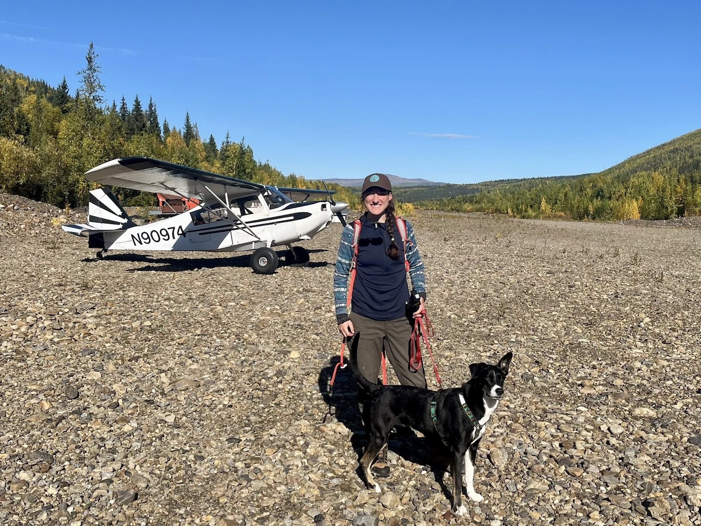 On a sunny fall day, a woman stands on a gravel bar with a black dog. A small airplane sits on the gravel bar in the background.