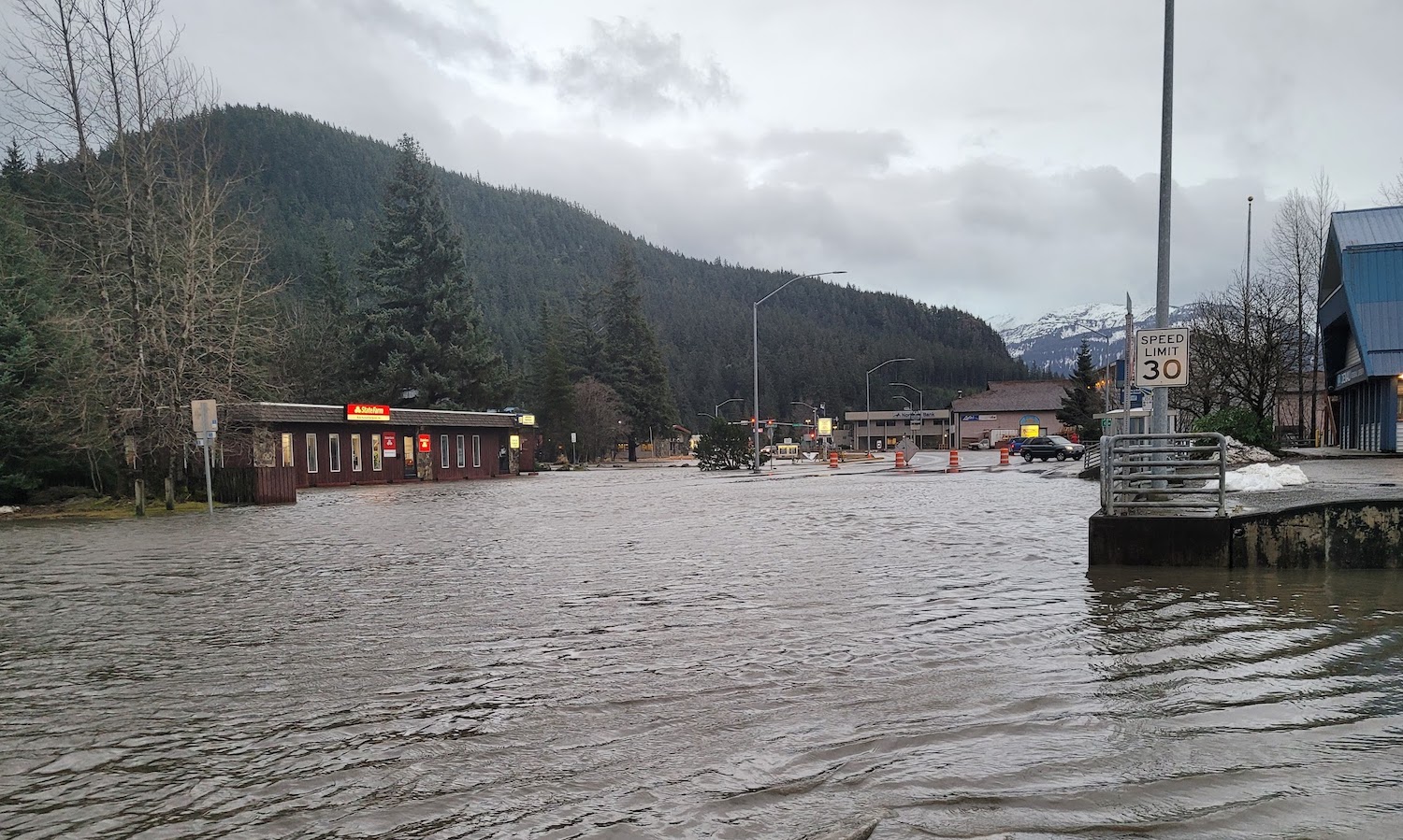 A pond covers a city street and adjacent business parking areas. Mountains covered with evergreens and snow form a backdrop under cloudy skies.