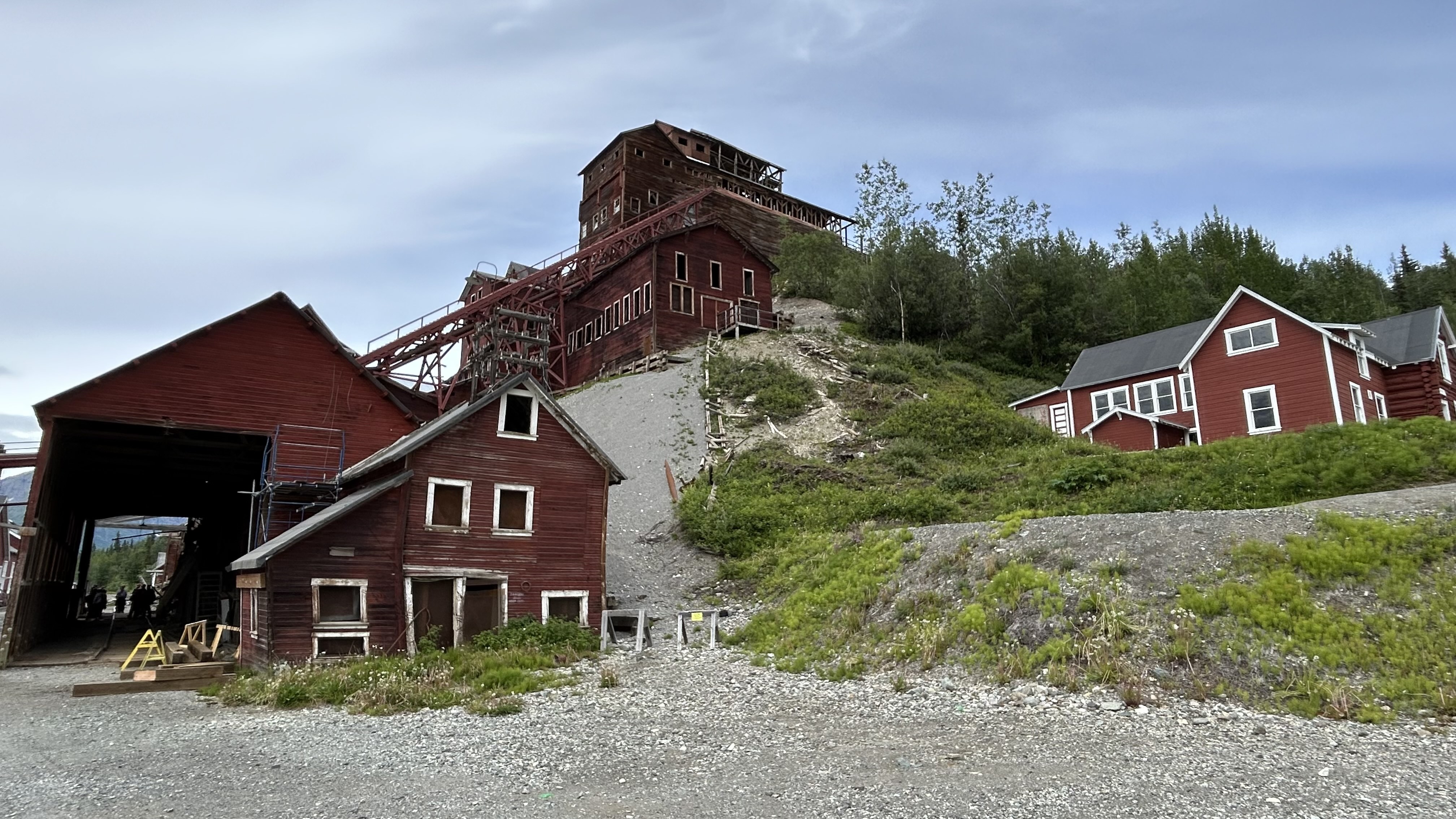 Historic red wooden buildings of the Kennecott Mines complex climb a green mountainside in Wrangell-St. Elias National Park and Preserve. The towering copper mill processed ore from the Bonanza, Jumbo, Mother Lode, Erie and Glacier mines; the restored mine office stands at right.
