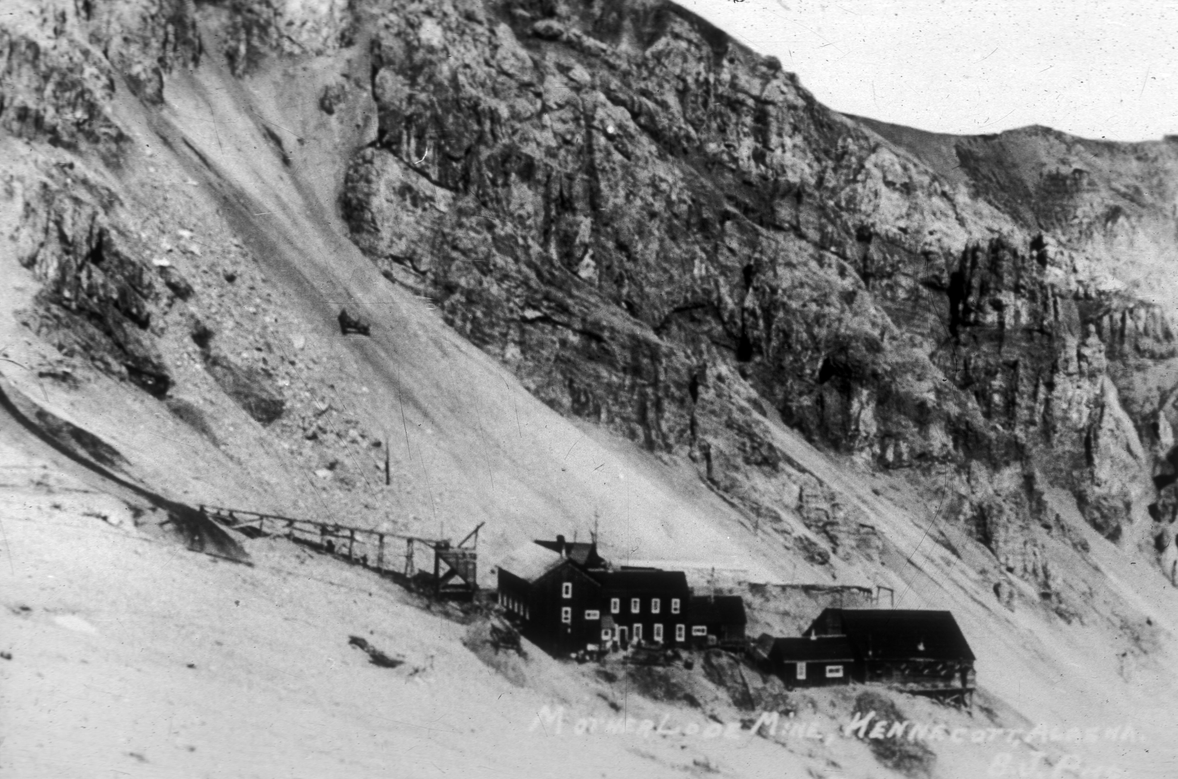 Black-and-white historic photograph of the Mother Lode Mine at Kennecott, Alaska, sometime between 1913 and 1939. Mine buildings sit on a steep, barren mountainside below towering cliffs, with elevated tramways and other mining structures extending across the slope.