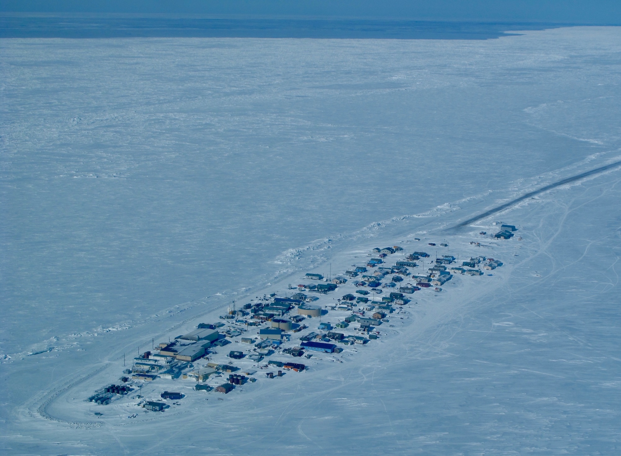 Viewed from an aircraft, a closely-packed group of buildings sit on a small spit of snow-covered land surrounded by sea ice. In the distance, beyond the ice, is an area of open water.