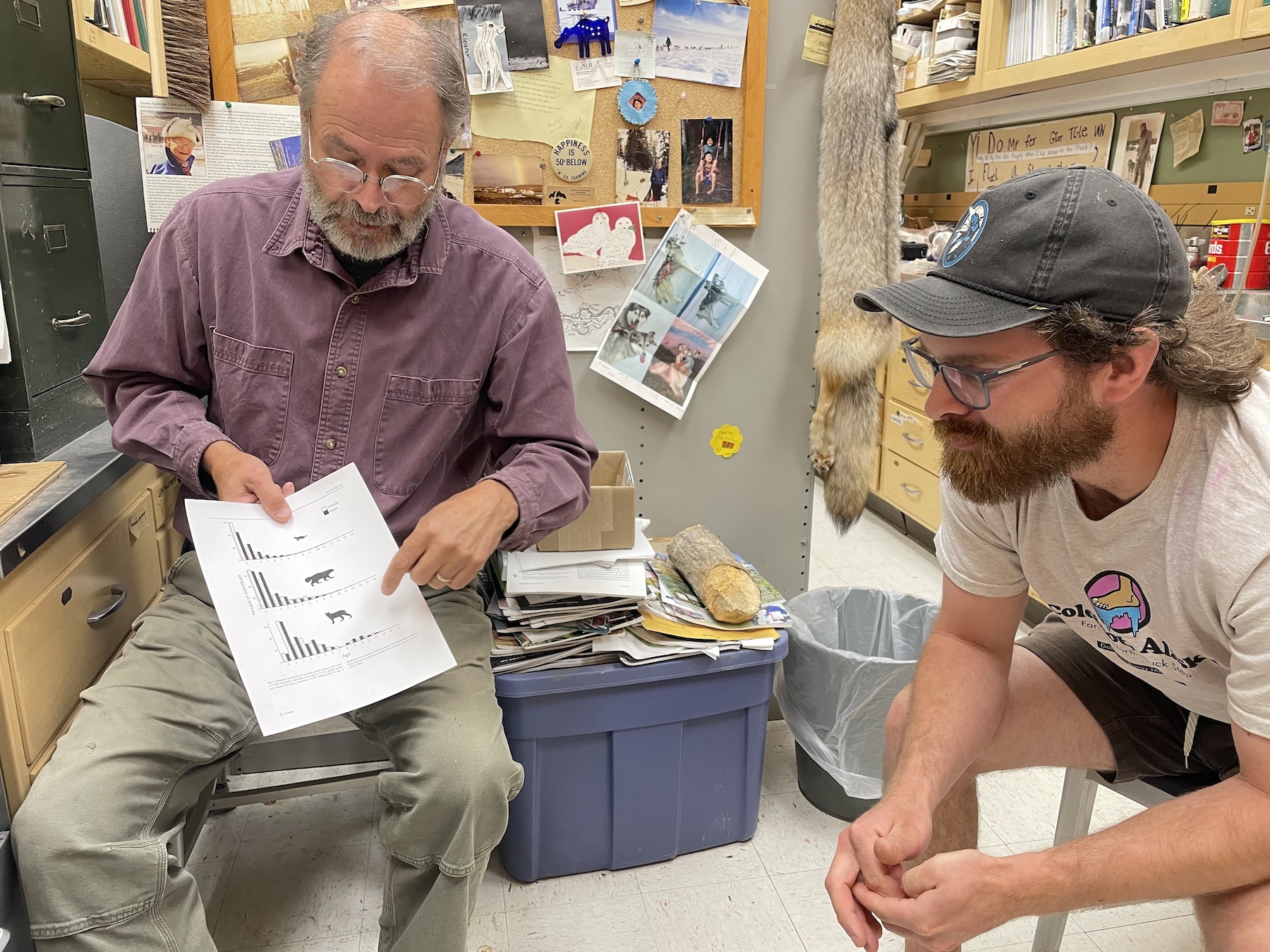 Two men sit in an office looking at a sheet of paper with graphs printed on it.