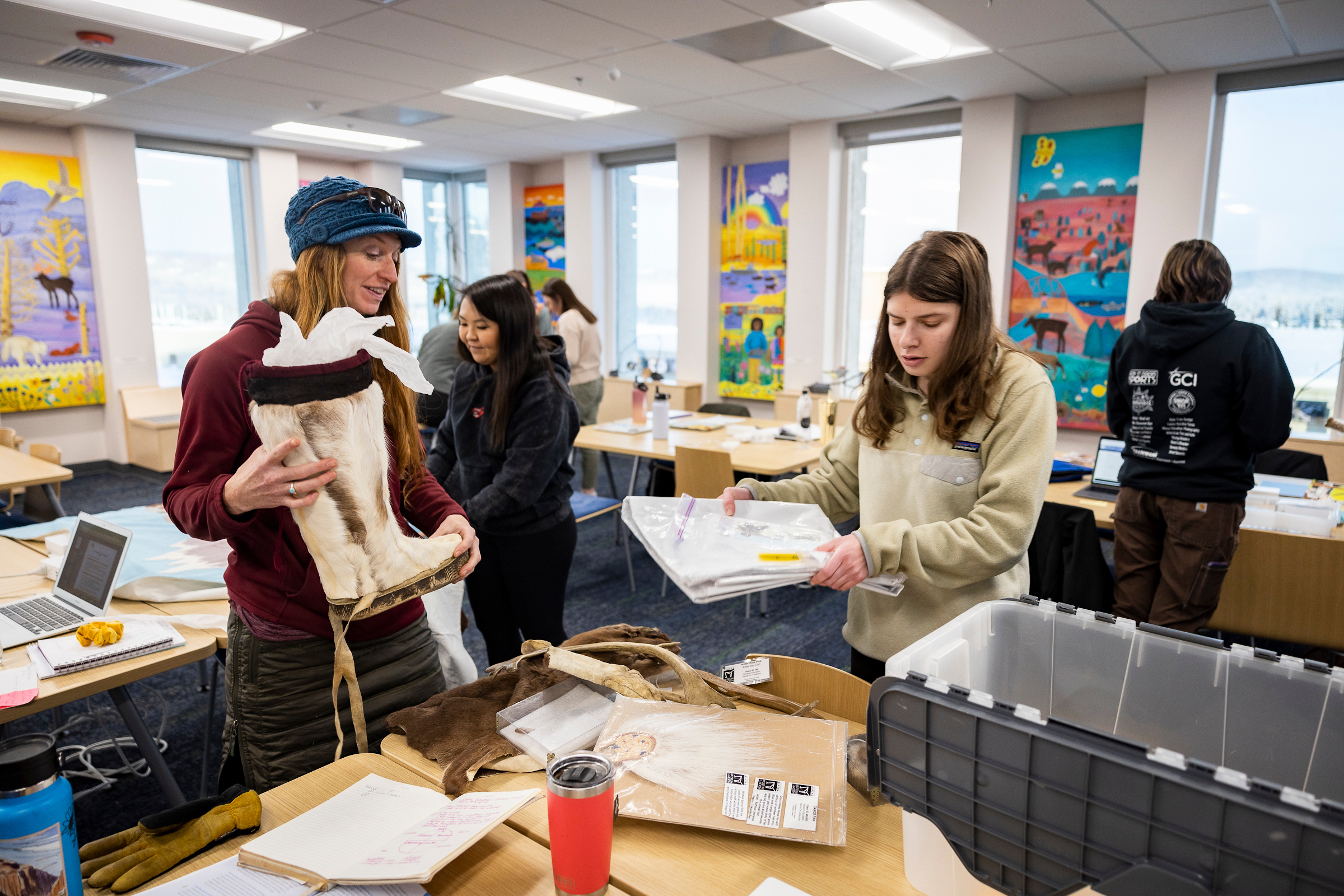 Five people standing in a classroom space with work tables and colorful posters on the walls. Two of them stand working together at a table: One holds a caribou hide mukluk and the other is lifting papers from a storage bin.