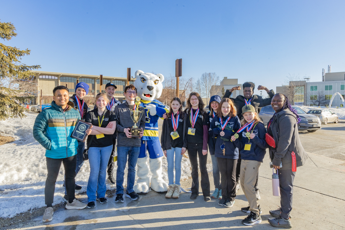 Ten middle school students wearing award medals pose with their two adult team coaches and the UAF Nook mascot outside in bright sun with a clear blue sky on the UAF campus. There is snow on the ground. In the background, there is the UAF Rasmuson library and engineering buildings, and the ice arch.